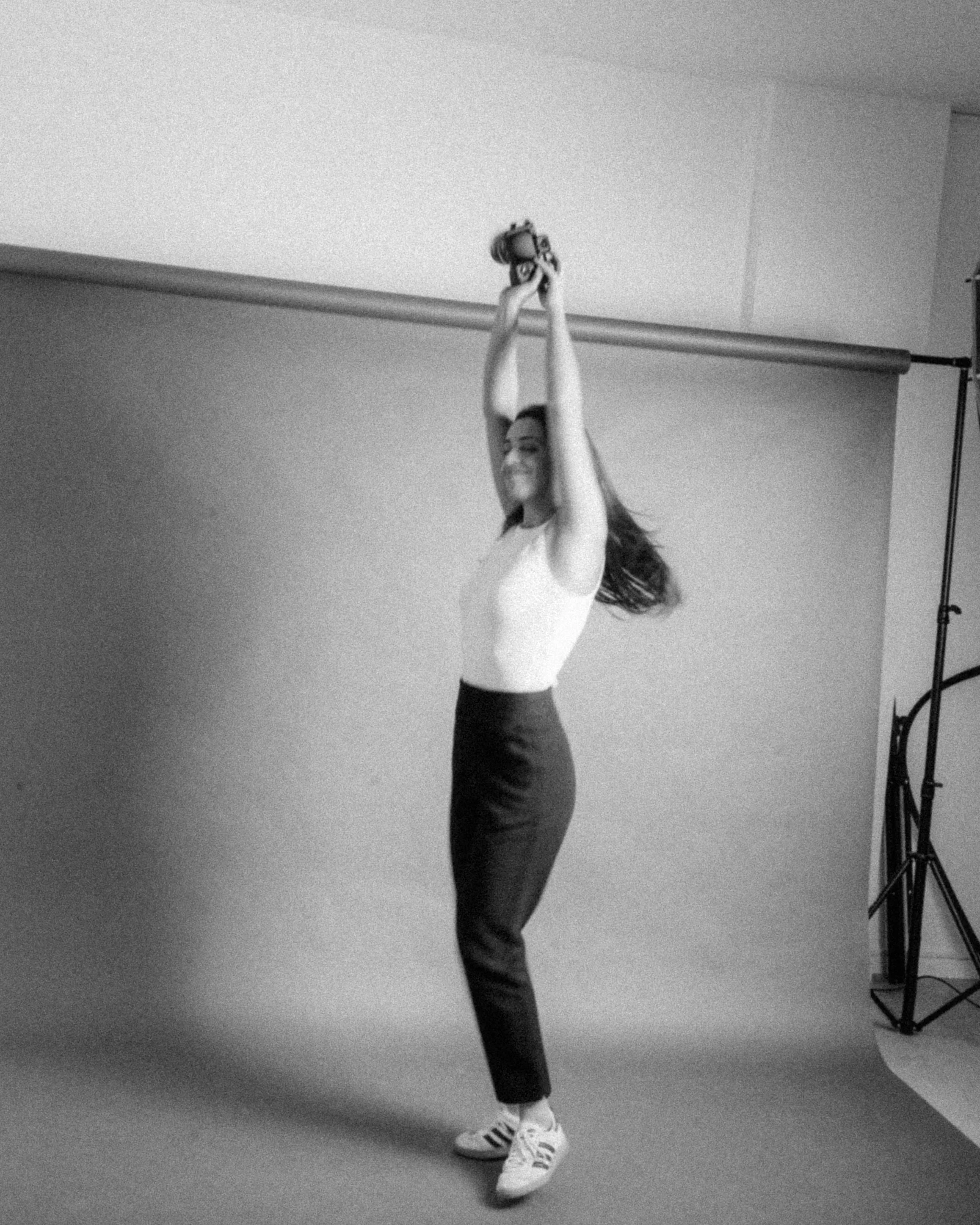 Videografa de casamentos a sorrir, com cabelo solto, a segurar uma câmera enquanto rodovia num estúdio, em preto e branco