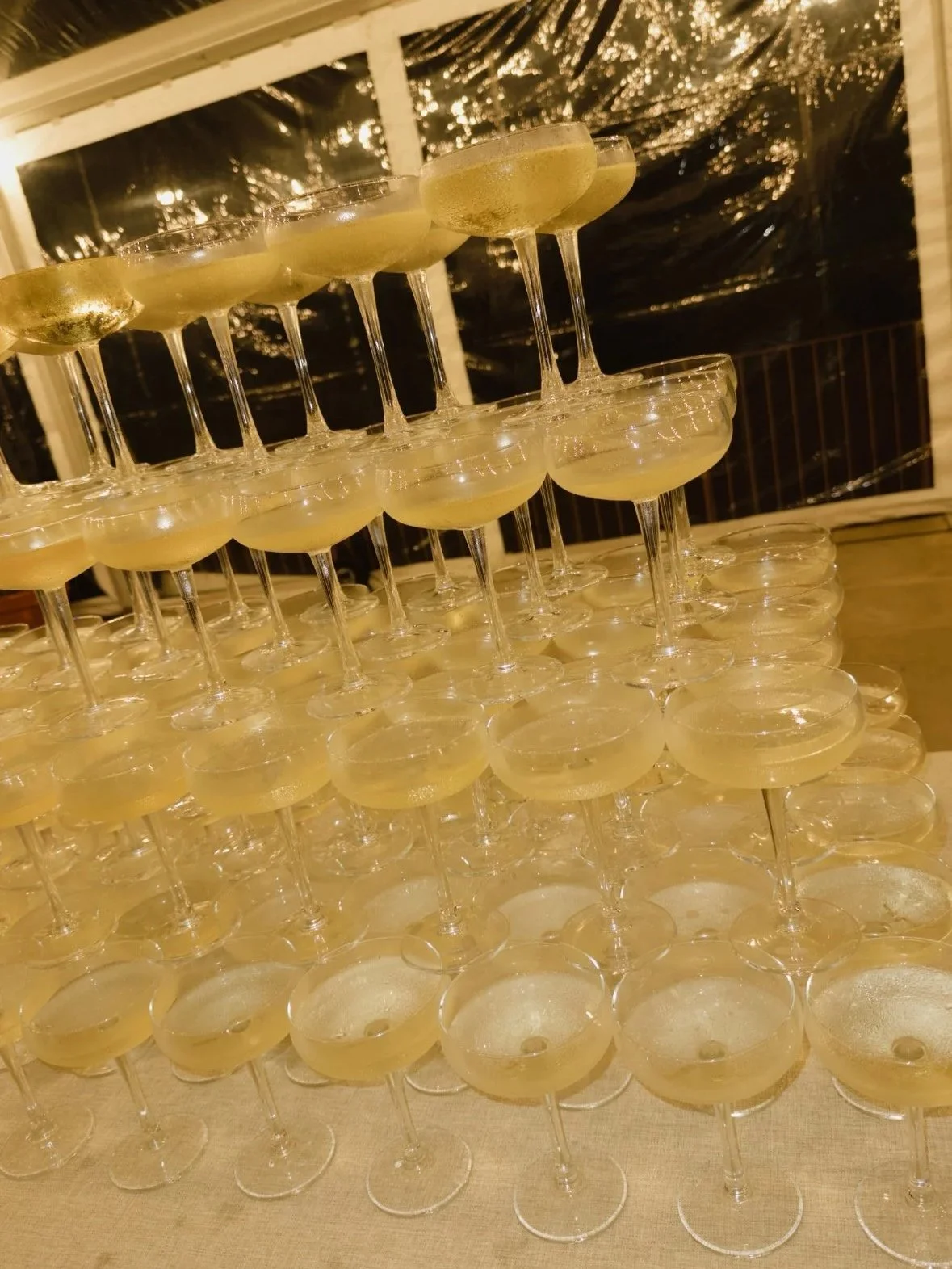 Várias taças de champanhe empilhadas em uma celebração, decoradas com luzes de festa ao fundo, no momento do corte do bolo de um casamento na quinta da abelheira em Mafra