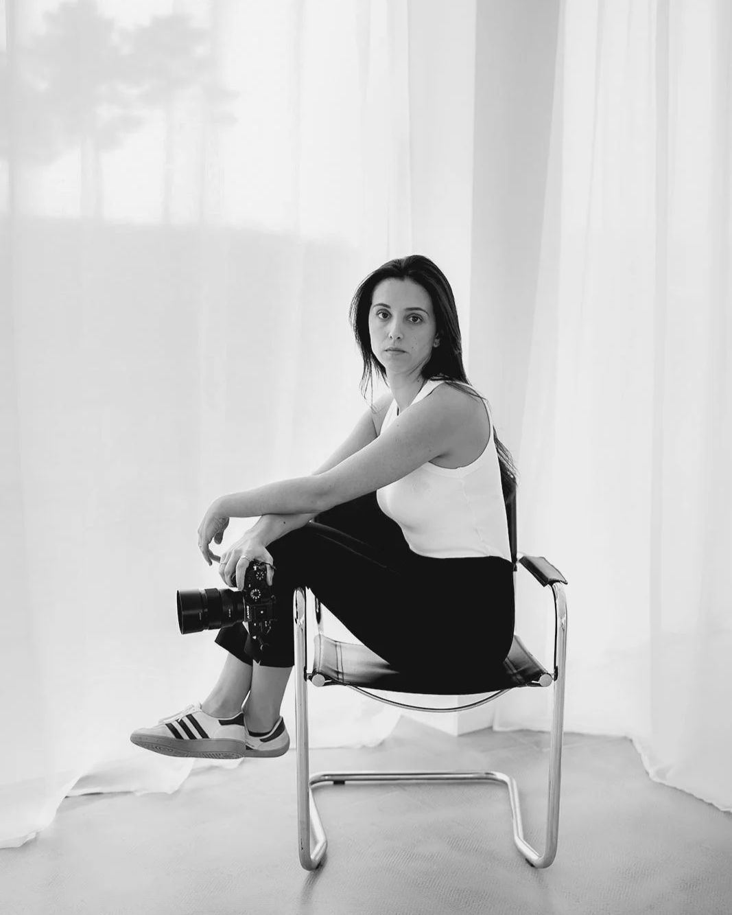 Videografa de casamentos sentada em uma cadeira, a segurar uma câmera, num ambiente com cortinas transparentes ao fundo, em preto e branco.