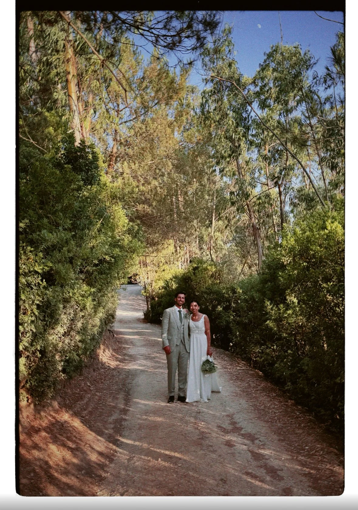 Casal de casamento caminhando por um caminho de terra rodeado por árvores verdes, sob céu azul com lua visível.