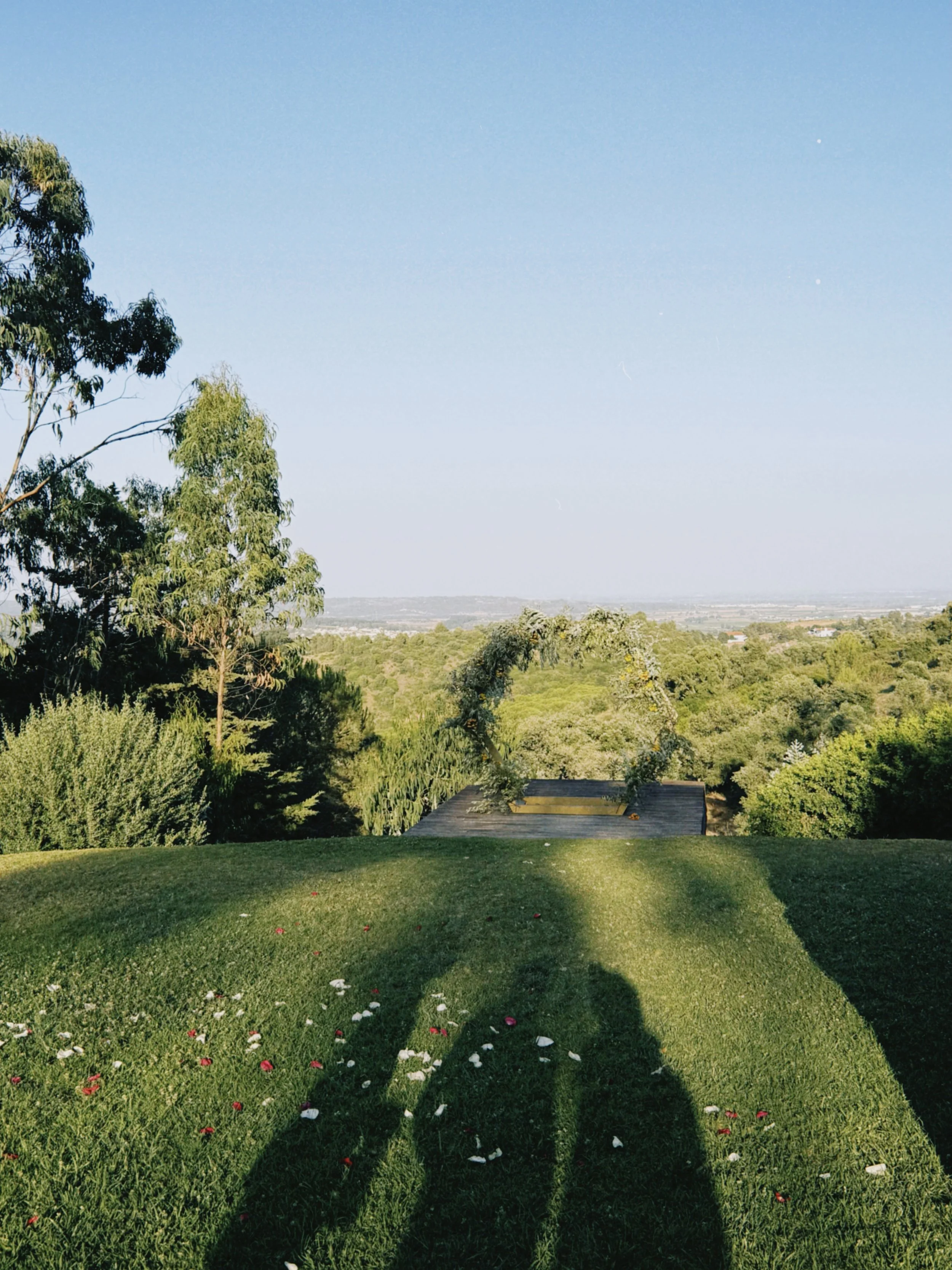 Paisagem com árvores, grama bem cuidada, flores espalhadas e sombras de duas pessoas na grama, com céu claro ao fundo.