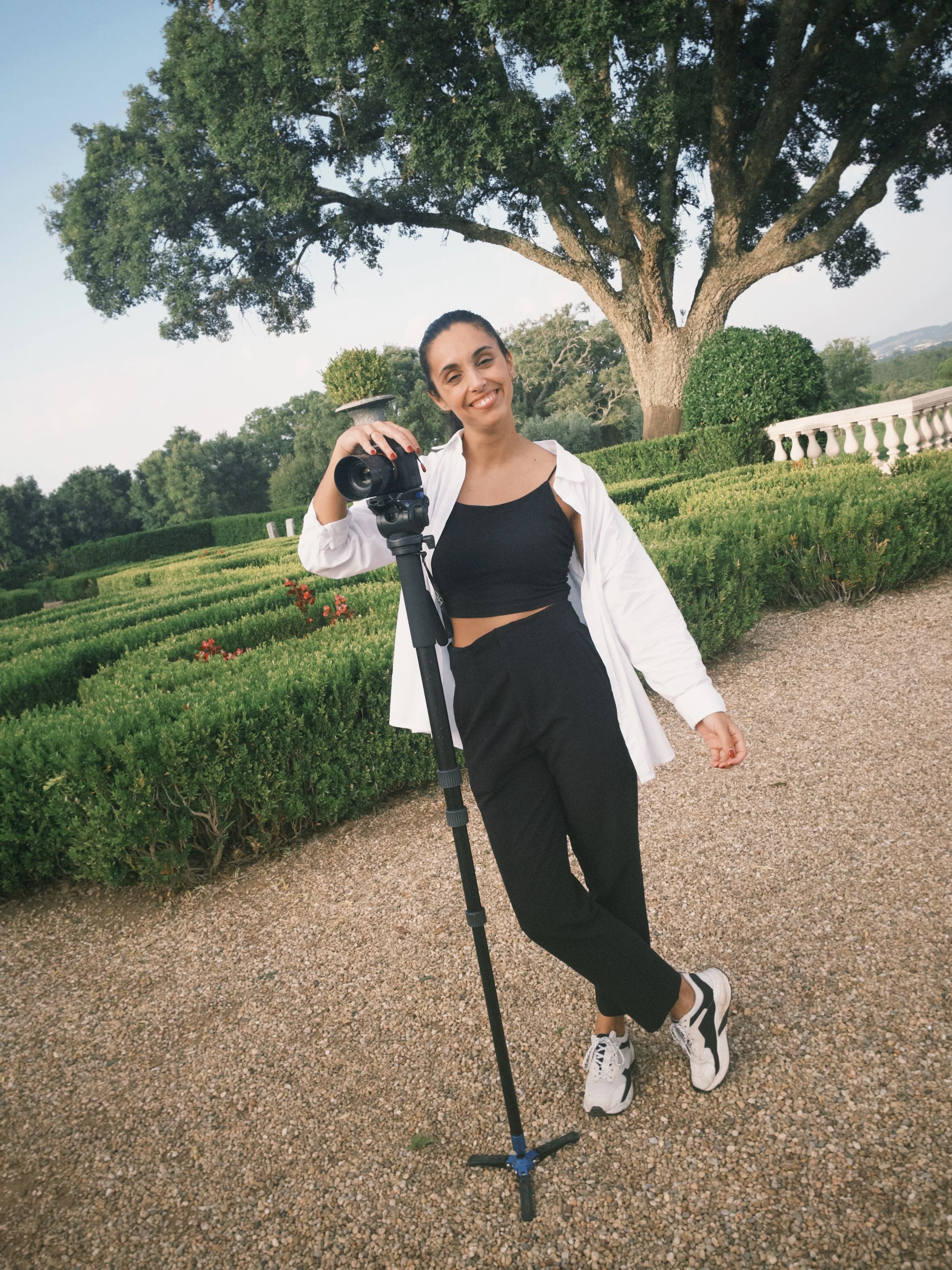 videografa mulher sorridente segurando um tripé com câmera, em um jardim com árvores e arbustos verdejantes ao fundo na herdade do peru
