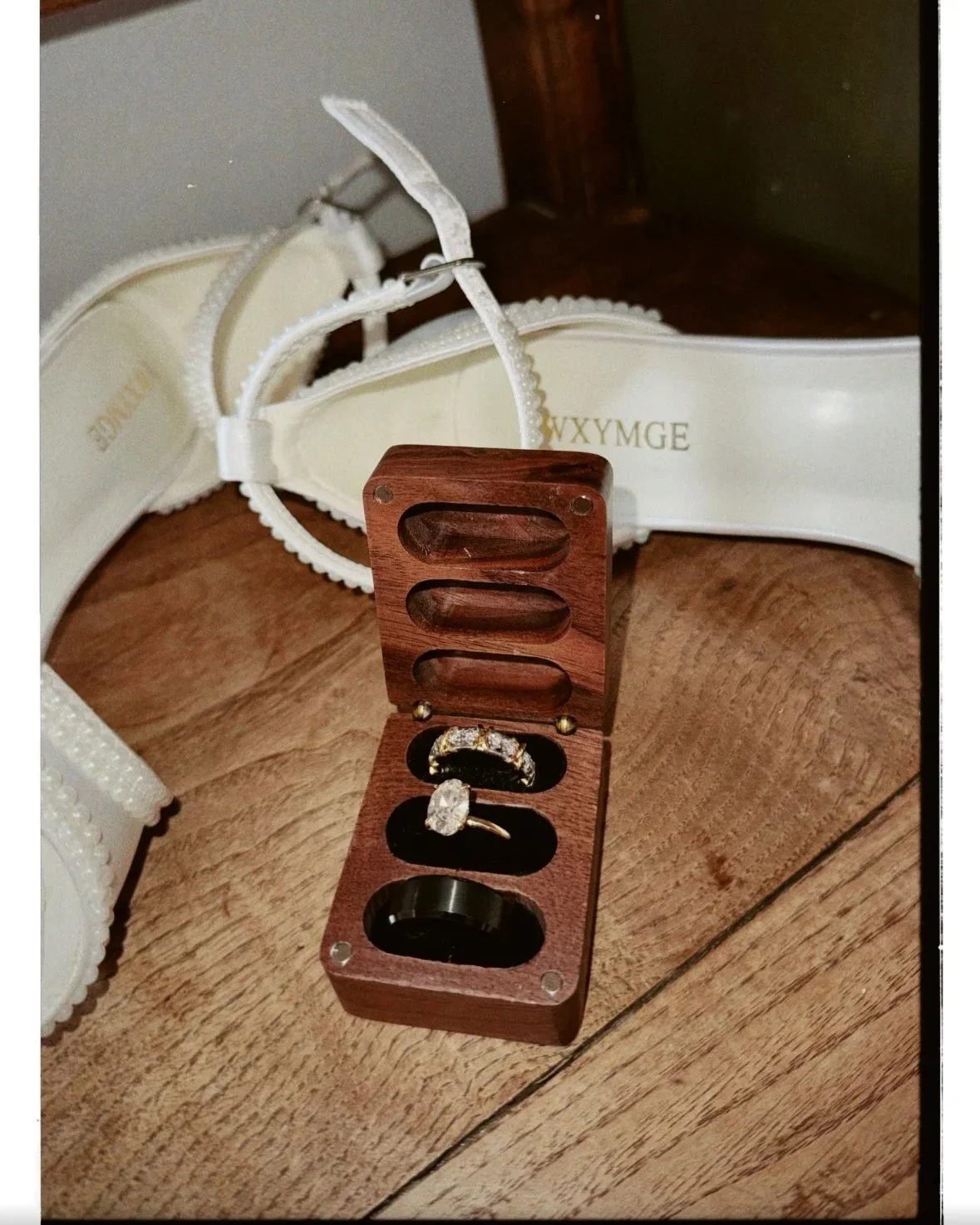 Caixa de madeira contendo três anéis de noivado, uma de prata com pedras brilhantes, uma de ouro e uma de metal escuro. Ao fundo, uma sandália branca de tiras com detalhes de pérolas e letras douradas 'WXYMGE'.