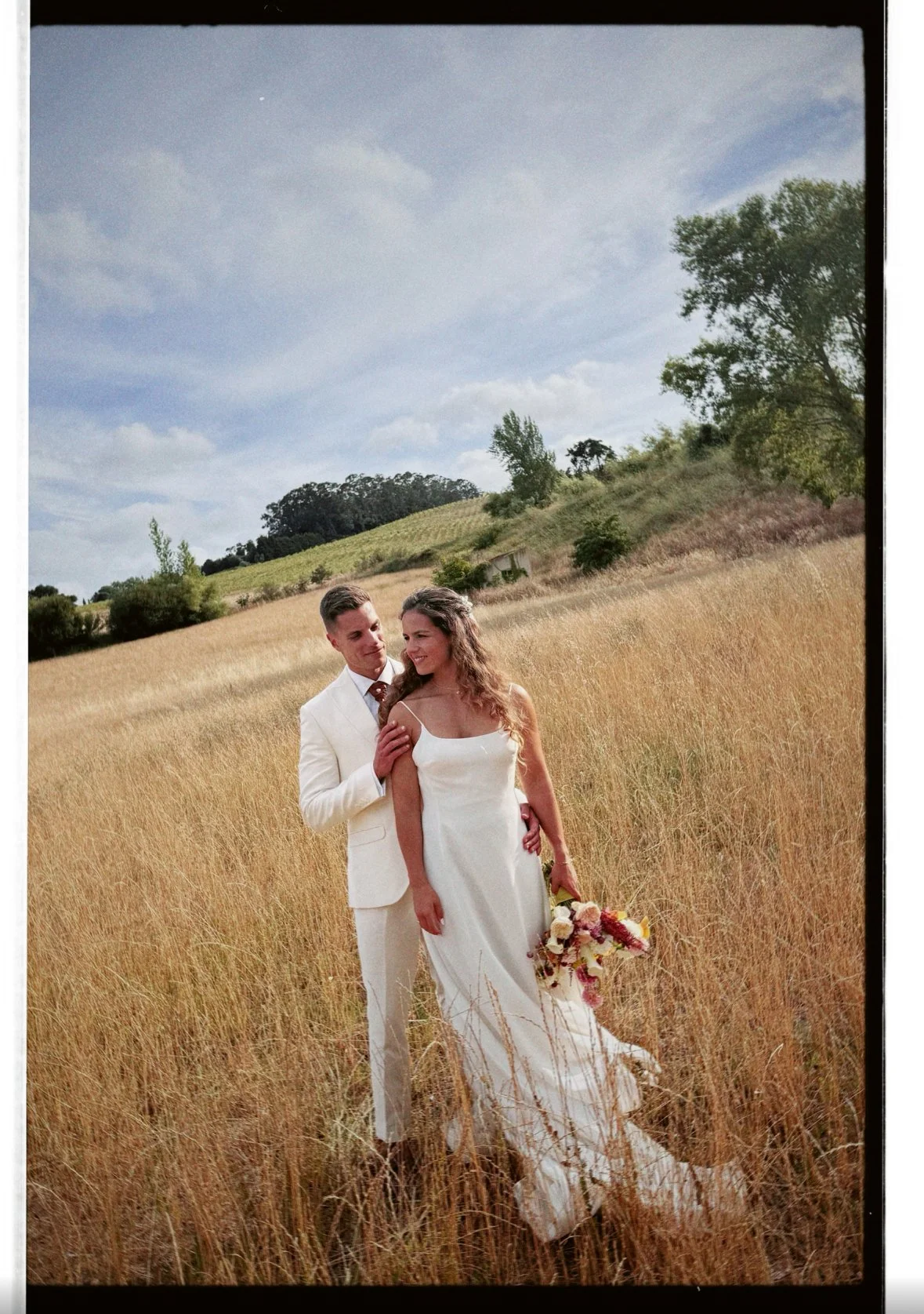 Noivos num campo de vegetação dourada com árvores e céu azul ao fundo em alenquer com vestido rosa clará