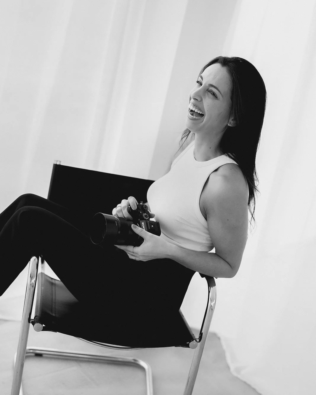 Videografa de casamentos sorridente de cabelo escuro a segurar uma câmera, sentada em uma cadeira de metal branco, em ambiente interno