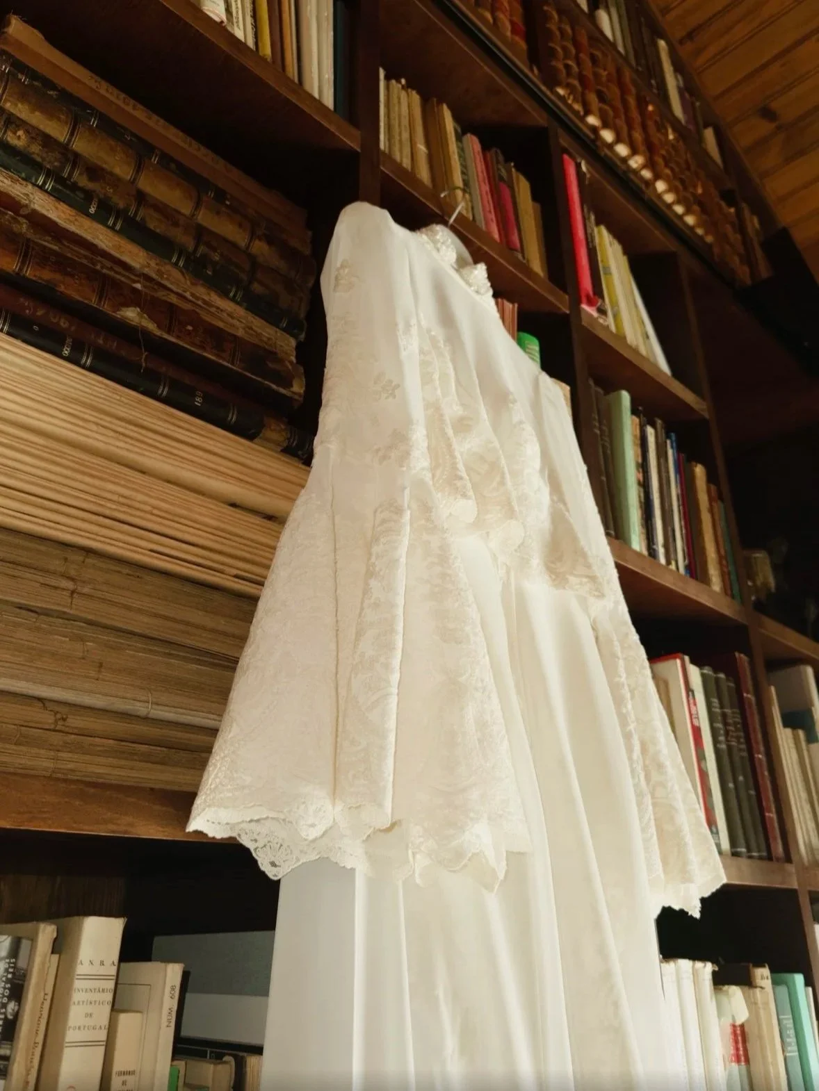 Vestido de noiva branco com detalhes em renda, pendurado em uma estante de livros de madeira na quinta da abelheira perto da ericeira