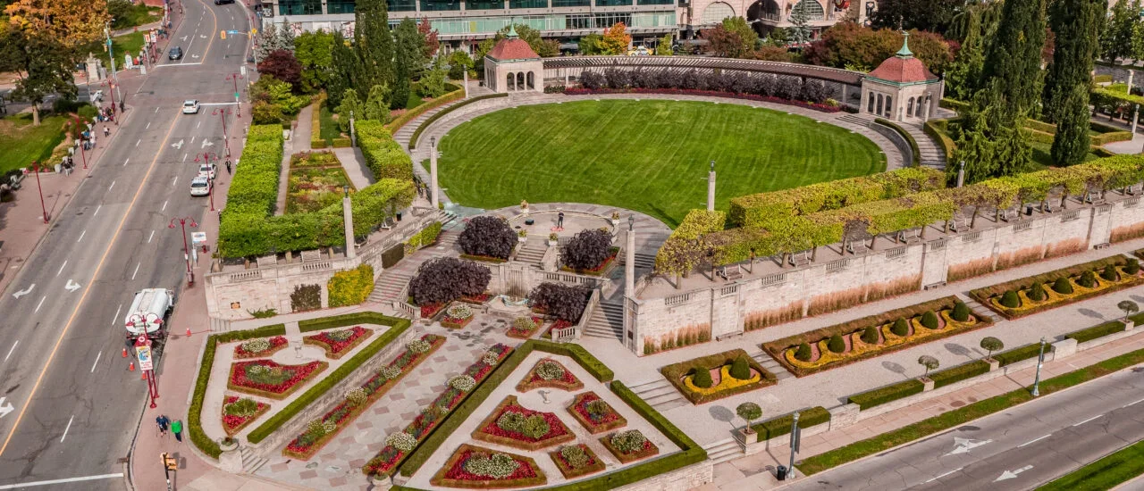 An aerial view of a city park with a maintained lawn, flowerbeds, paved walkways, trees, and small pavilions, bordered by streets with cars and pedestrians.