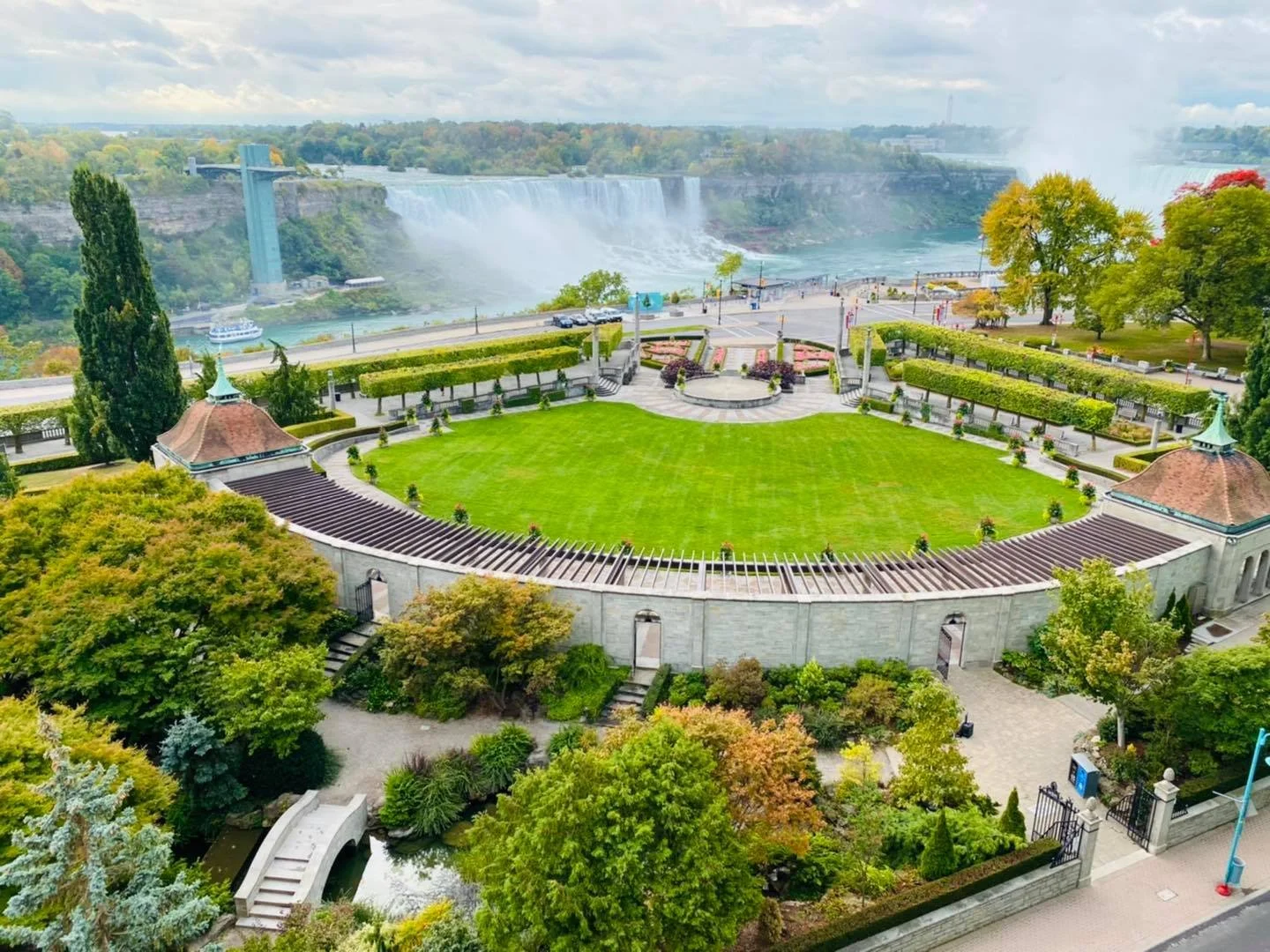 A large park with a central green lawn, surrounded by trees and pathways, overlooking Niagara Falls in the distance.