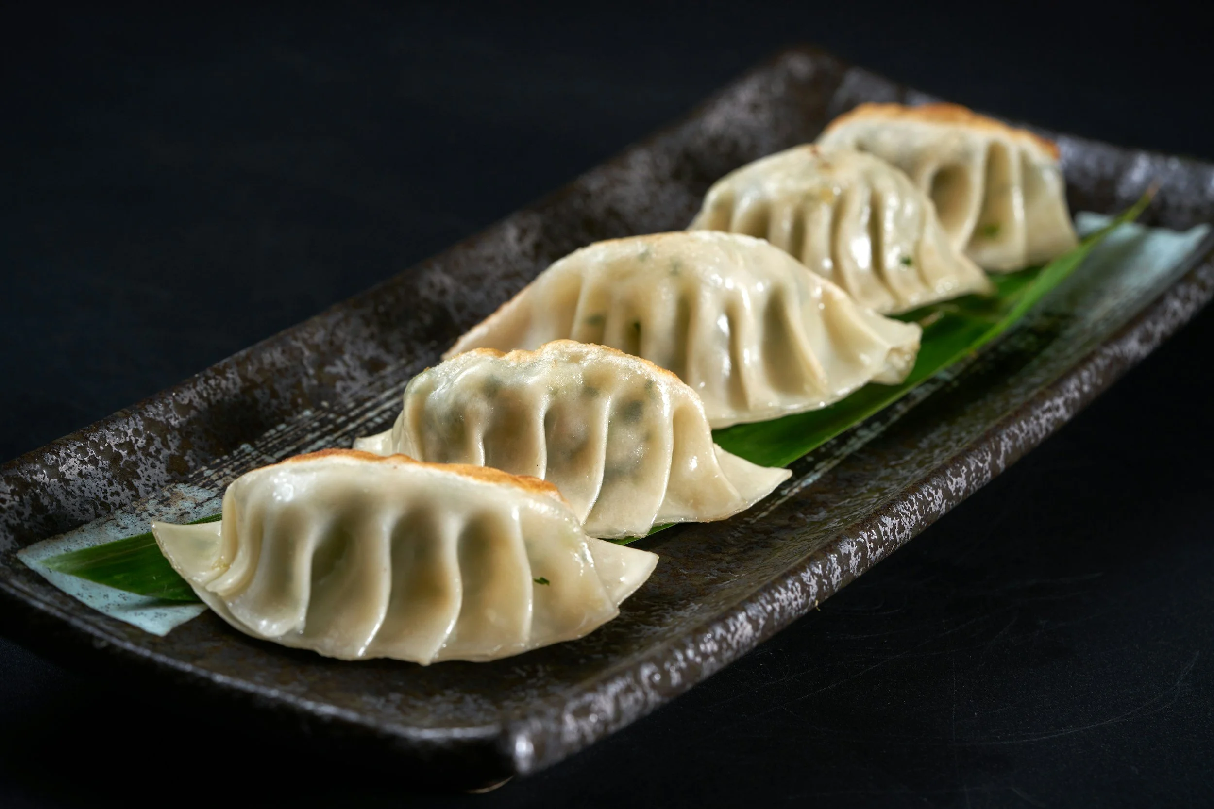 Close-up of five steamed dumplings arranged on a black ceramic plate with a green leaf underneath, against a dark background.
