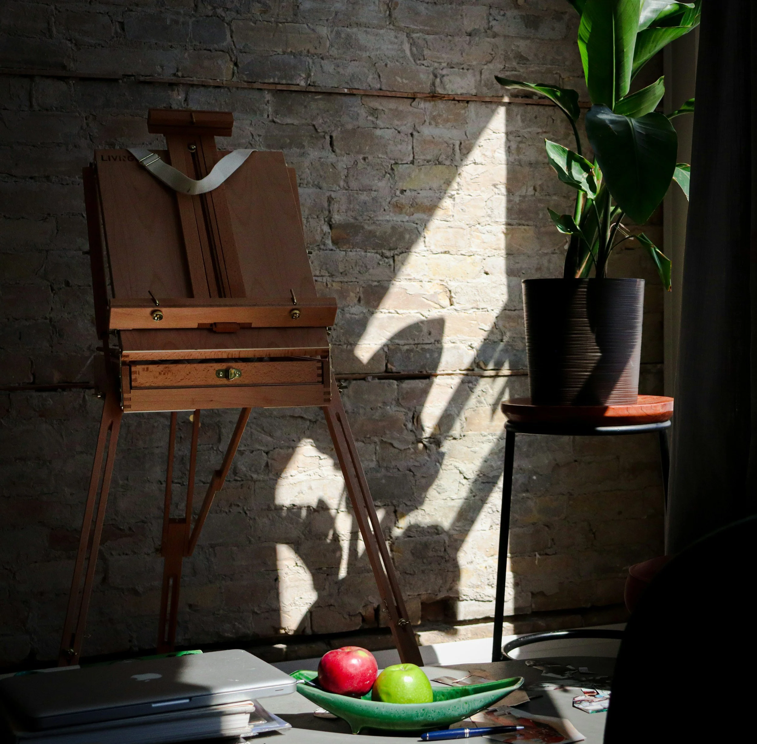 A workspace with a variety of items including apples in a green dish, a laptop, books, and papers on a table. In the background, there is a brick wall, a large green plant, and a light sunlight casting shadows.