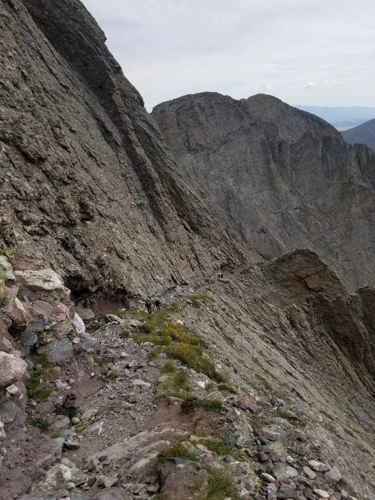 Three hikers traversing on a narrow mountain trail surrounded by steep rocky slopes and rugged cliffs.
