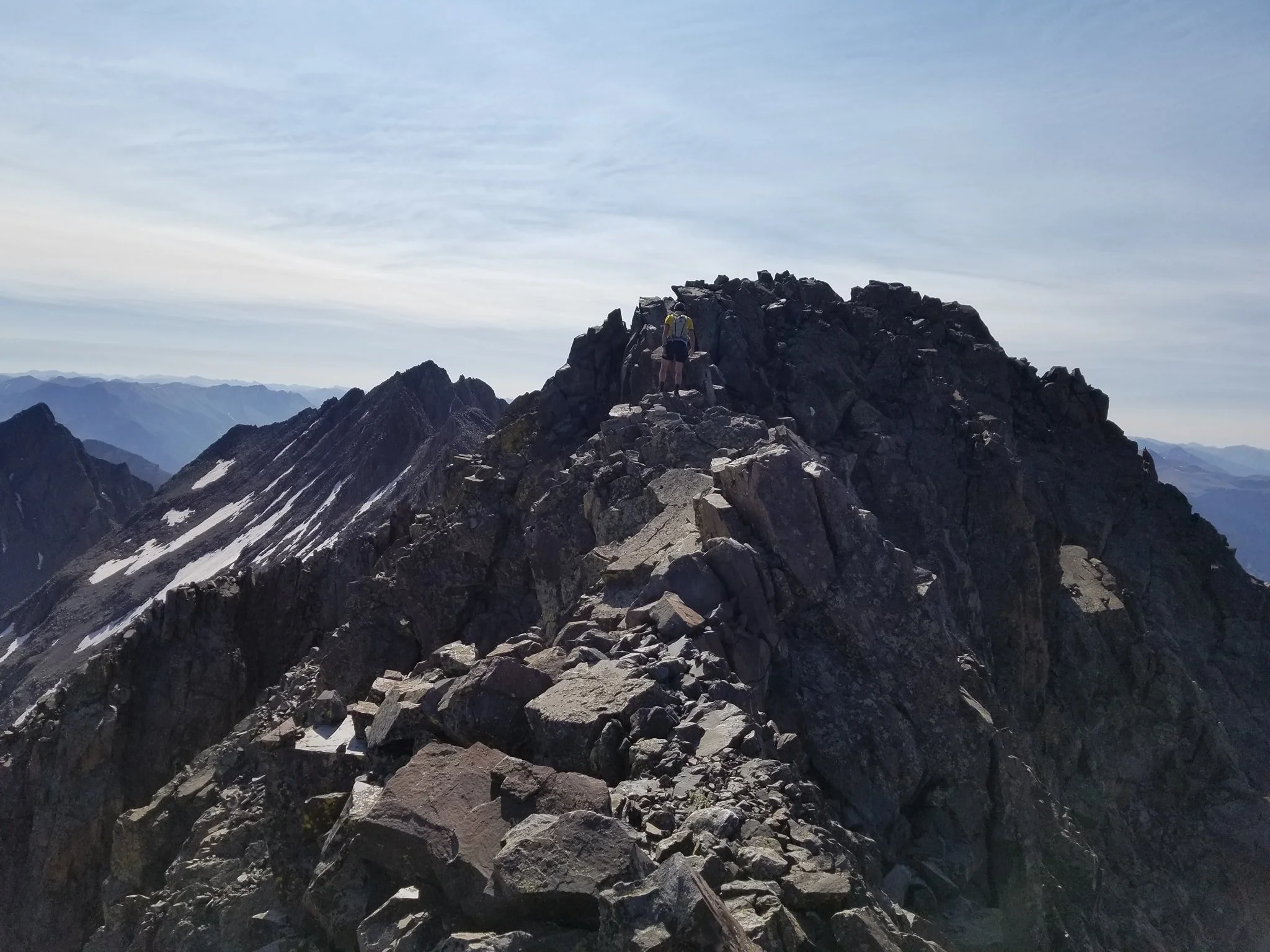 Hikers climbing a rocky mountain peak with snow patches, under a cloudy sky, with distant mountain ridges in the background.