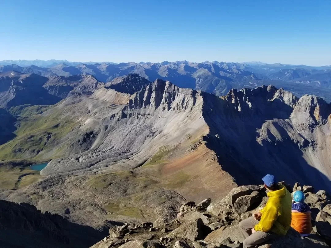 Two hikers sitting on rocks overlooking a vast mountain range with rugged peaks, a small lake, and clear blue sky.