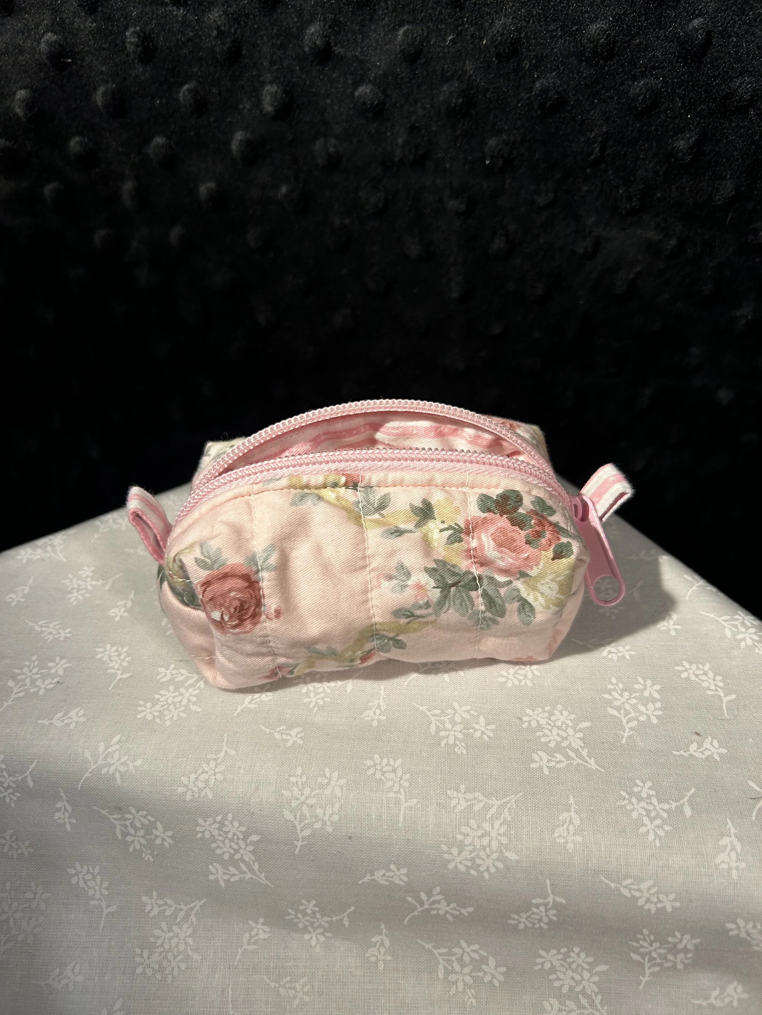 A small pink floral-patterned zippered pouch sitting on a white fabric surface with a subtle floral design.