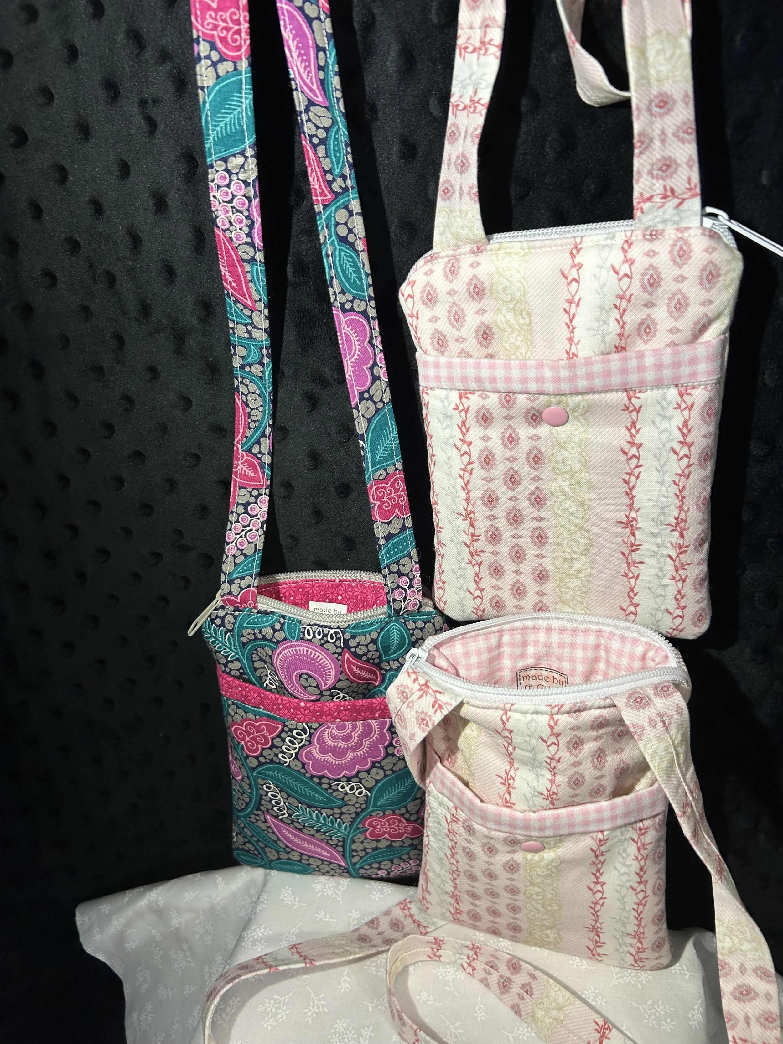 A colorful fabric shoulder bag with a paisley and floral pattern in black, pink, teal, and purple, alongside two smaller matching pouches with pink and cream patterns, all displayed against a black textured background.