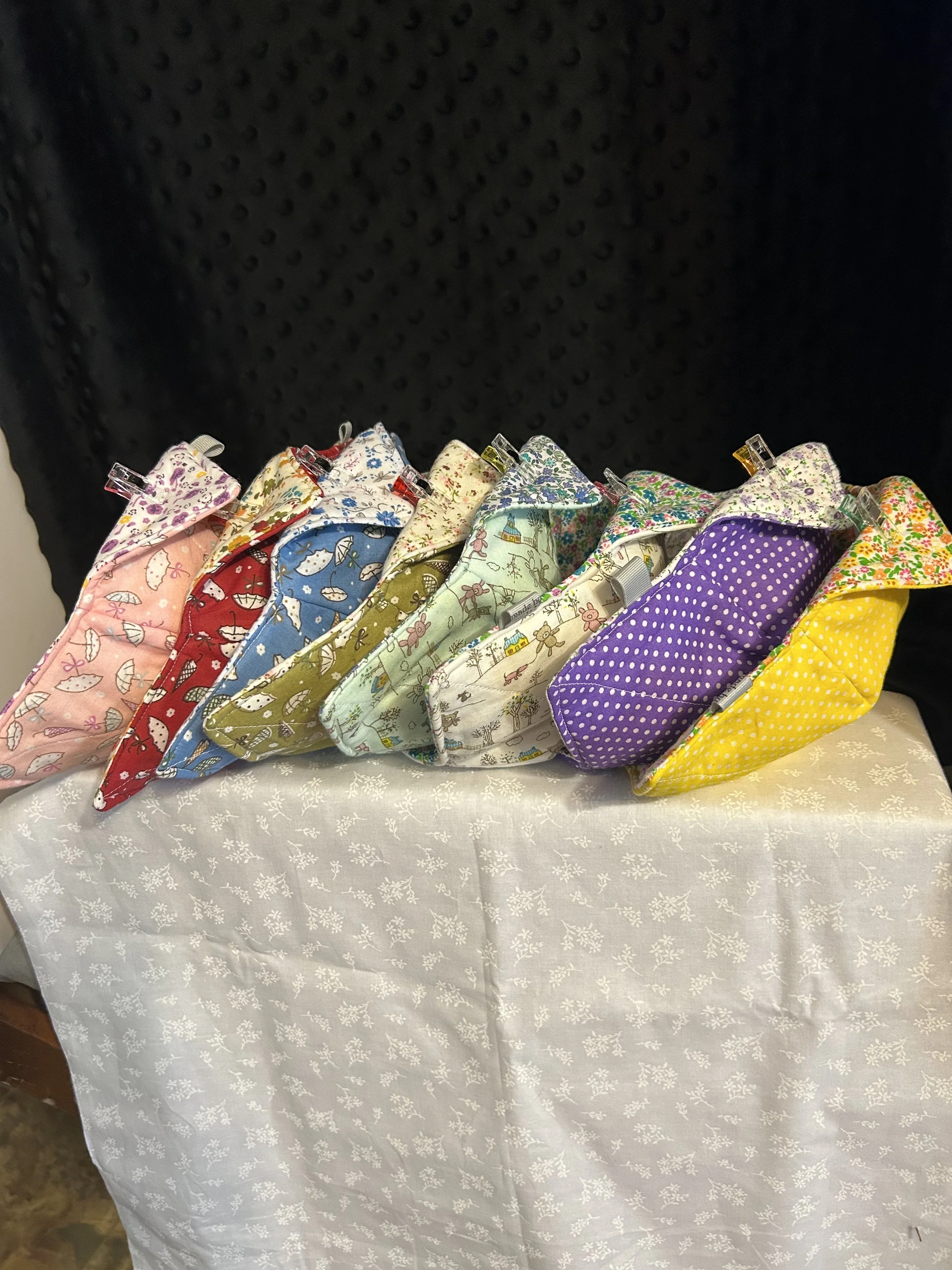A display of colorful handmade fabric face masks arranged on a table covered with a white cloth with a snowflake pattern, set against a black background.