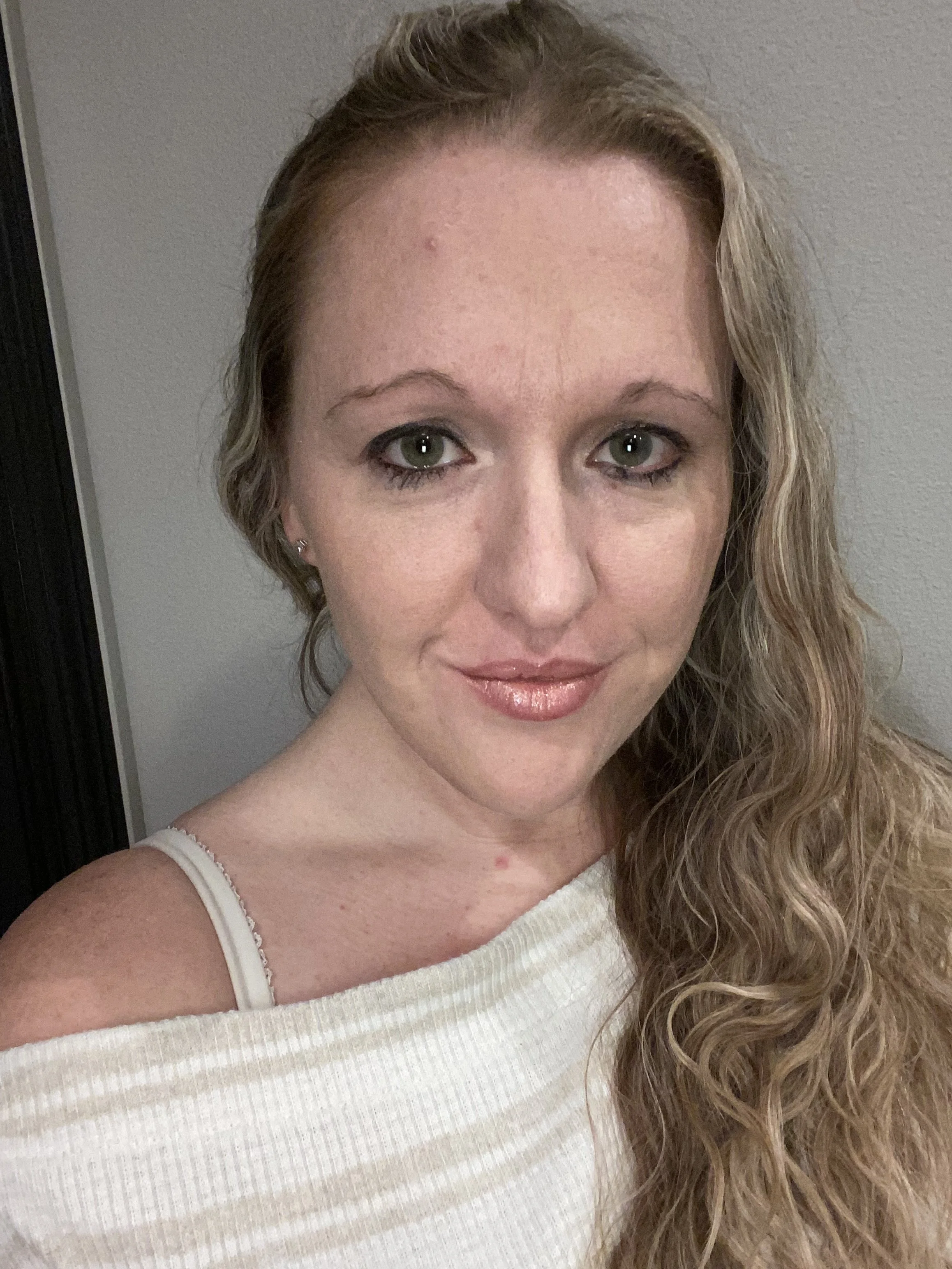 Close-up portrait of a woman with long, wavy blonde hair, light green eyes, and fair skin, wearing a cream-colored off-the-shoulder top and a white bra strap visible on her left shoulder.