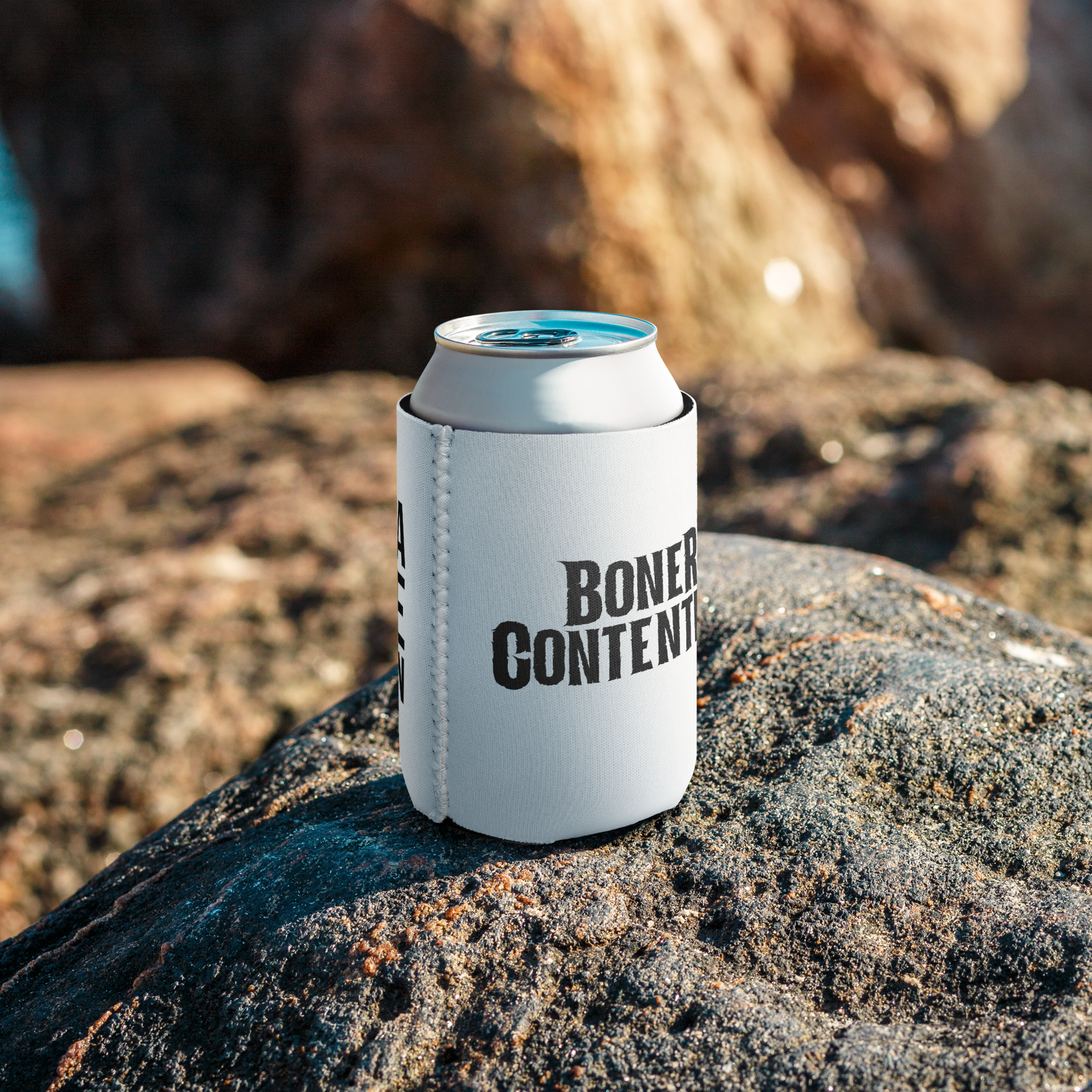 A white can with a black label that says "Boner Contention" is sitting on a rough, dark rock with small glistening grains. The background is blurred with warm tones and sunlight.