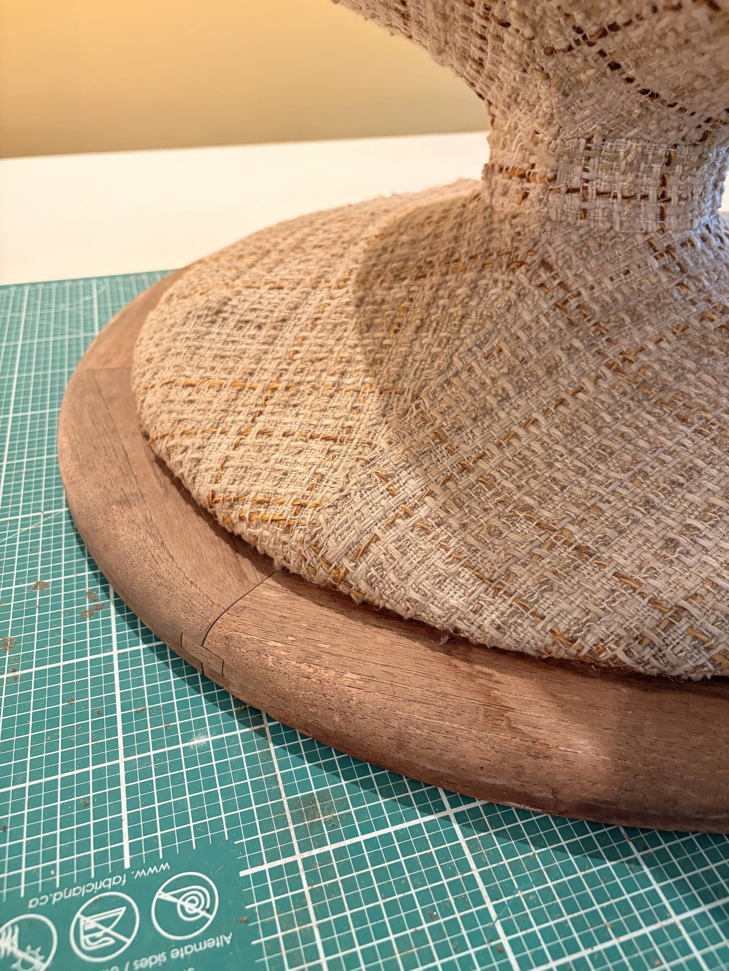 Close-up of a woven fabric hat on a wooden display stand, placed on a green cutting mat with grid lines. Need is in need of refinishing