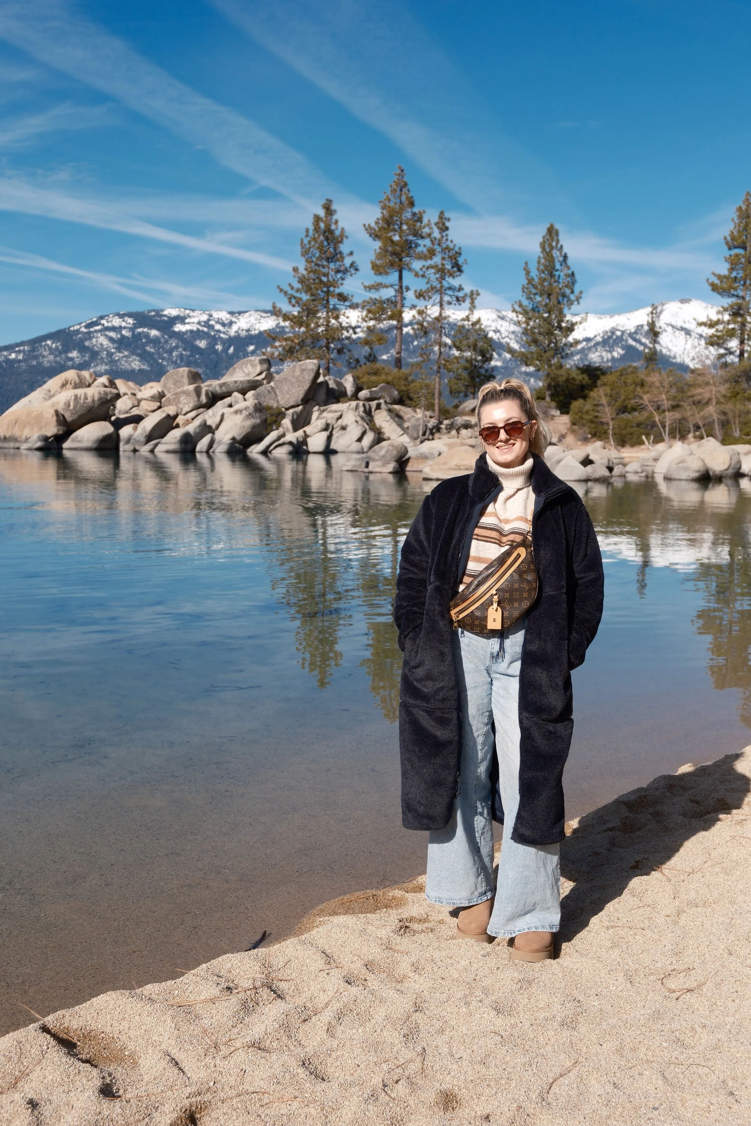 A woman standing on sandy shore next to a lake with rocky shoreline, pine trees, snowy mountains, and blue sky in the background.