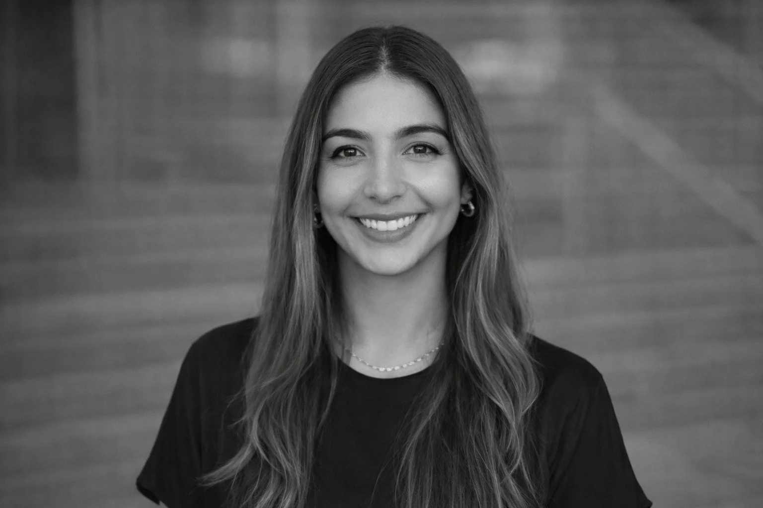 Retrato de una mujer joven sonriendo, con cabello largo y ondulado, usando un collar y pendientes, contra un fondo de madera.