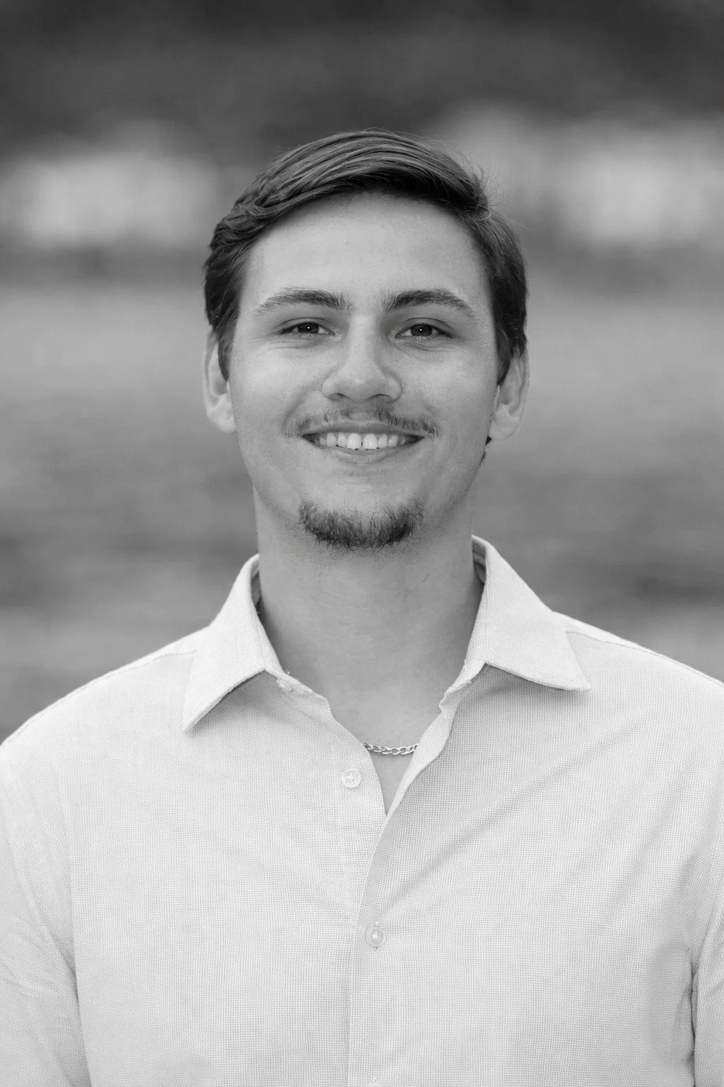 Retrato en blanco y negro de un joven sonriendo, lleva una camisa y un cadena, fondo blurred de paisaje natural.