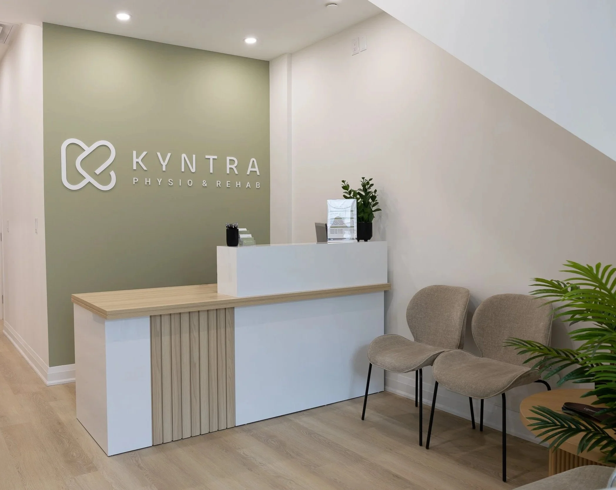 Reception desk in a modern interior with a sign reading "Kyntra Physio & Rehab" on a green wall, chairs, plants, and a clear acrylic stand on the desk.