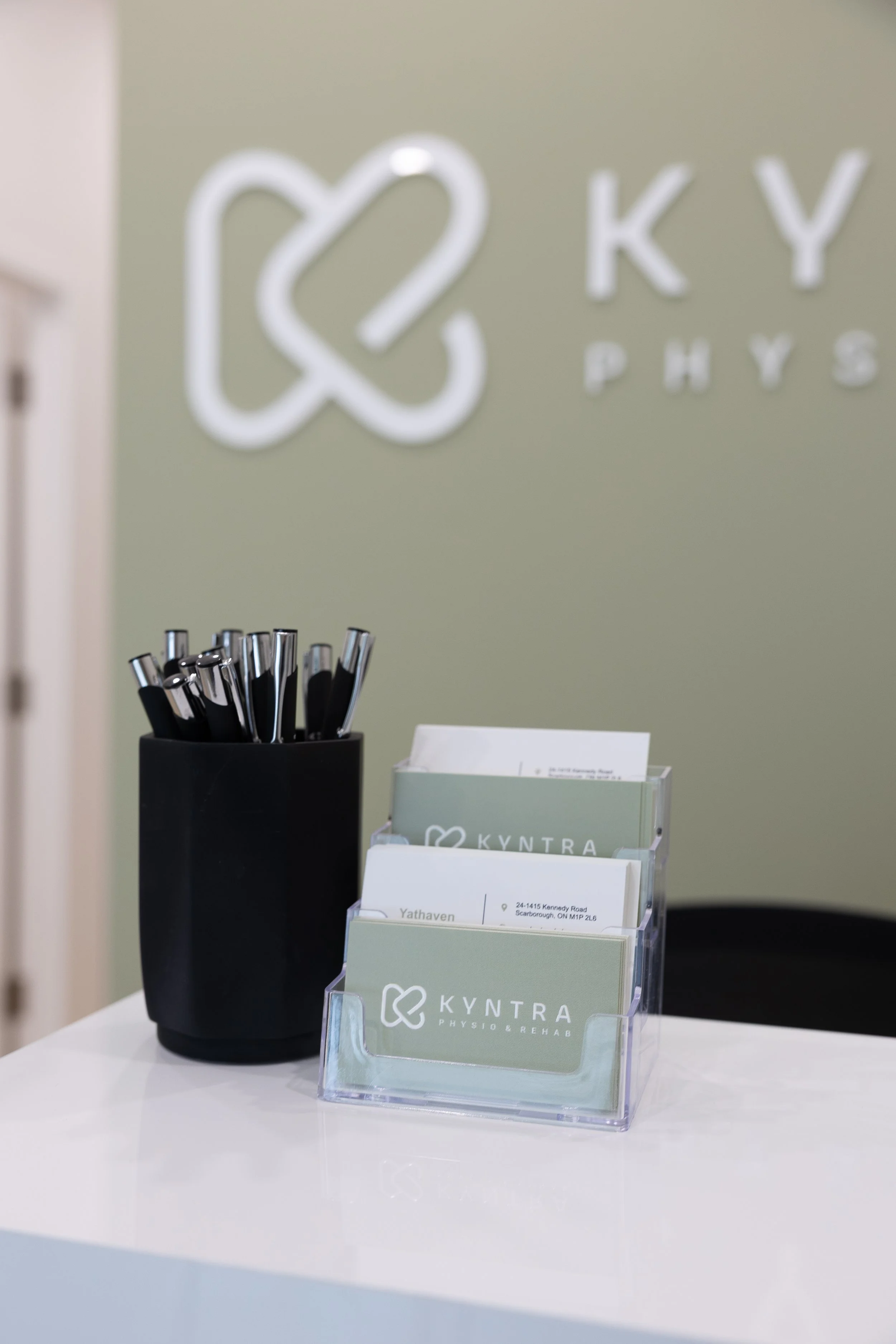 Business reception area with a black pen holder with pens and a clear organization holder with business cards on a white desk, with a gray wall in the background featuring a white logo and text.