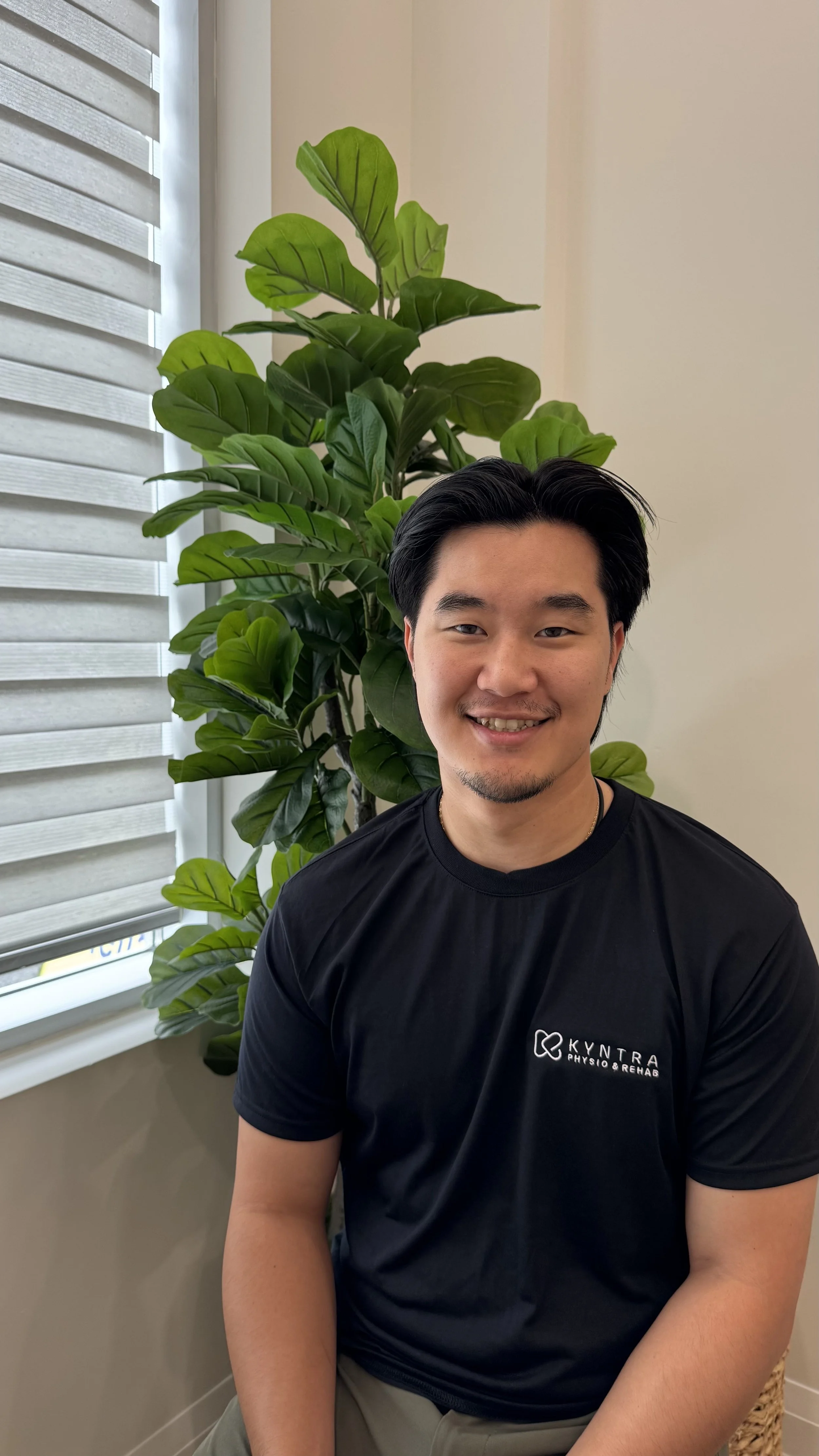 A smiling young man with black hair and light skin in a black shirt sitting in front of a large green leafy plant near a window with white blinds.