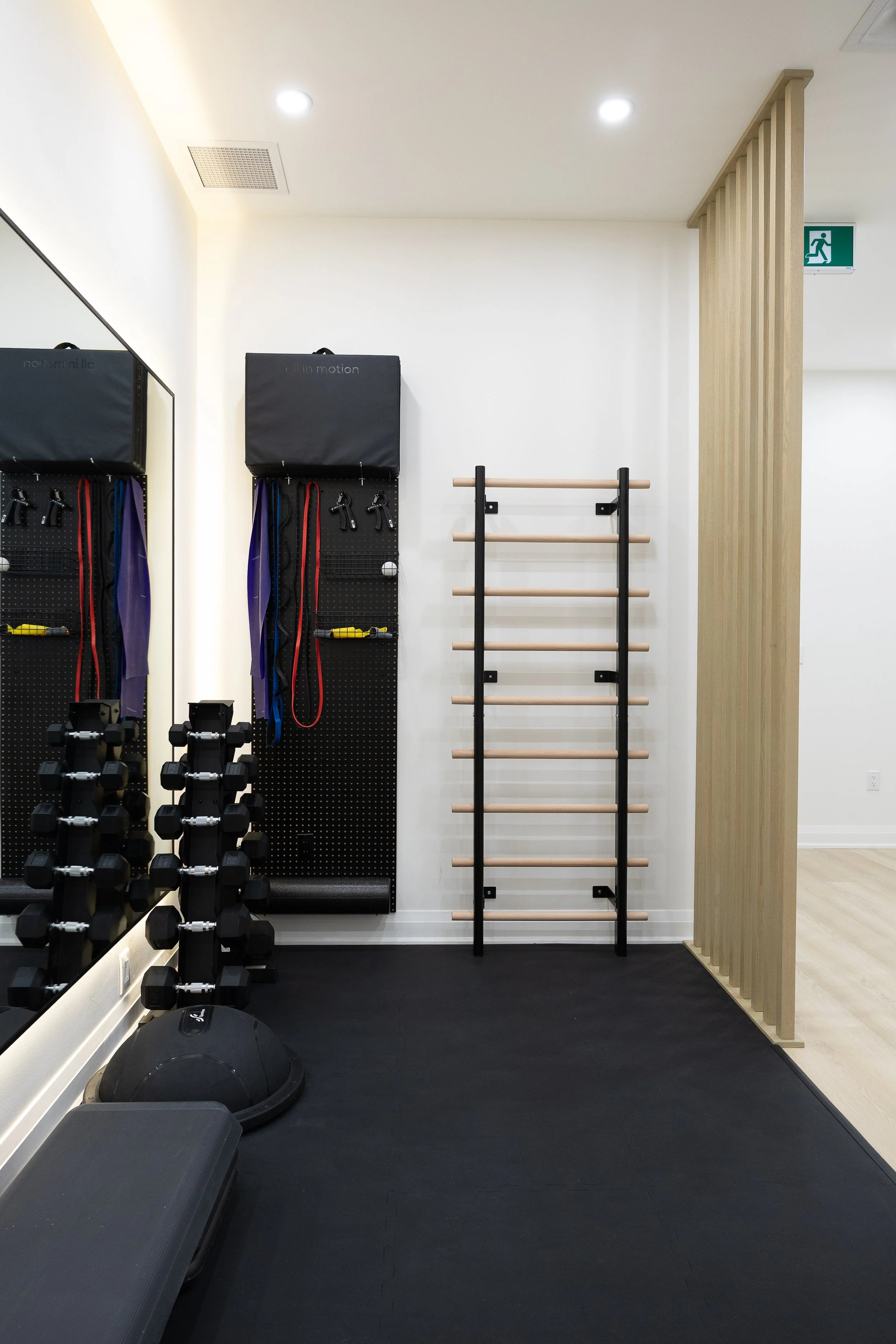 A gym corner with black dumbbell rack, medicine balls, jump ropes, padded mats, a wooden ladder, and an emergency exit sign. The space has white walls, ceiling lights, and black flooring.