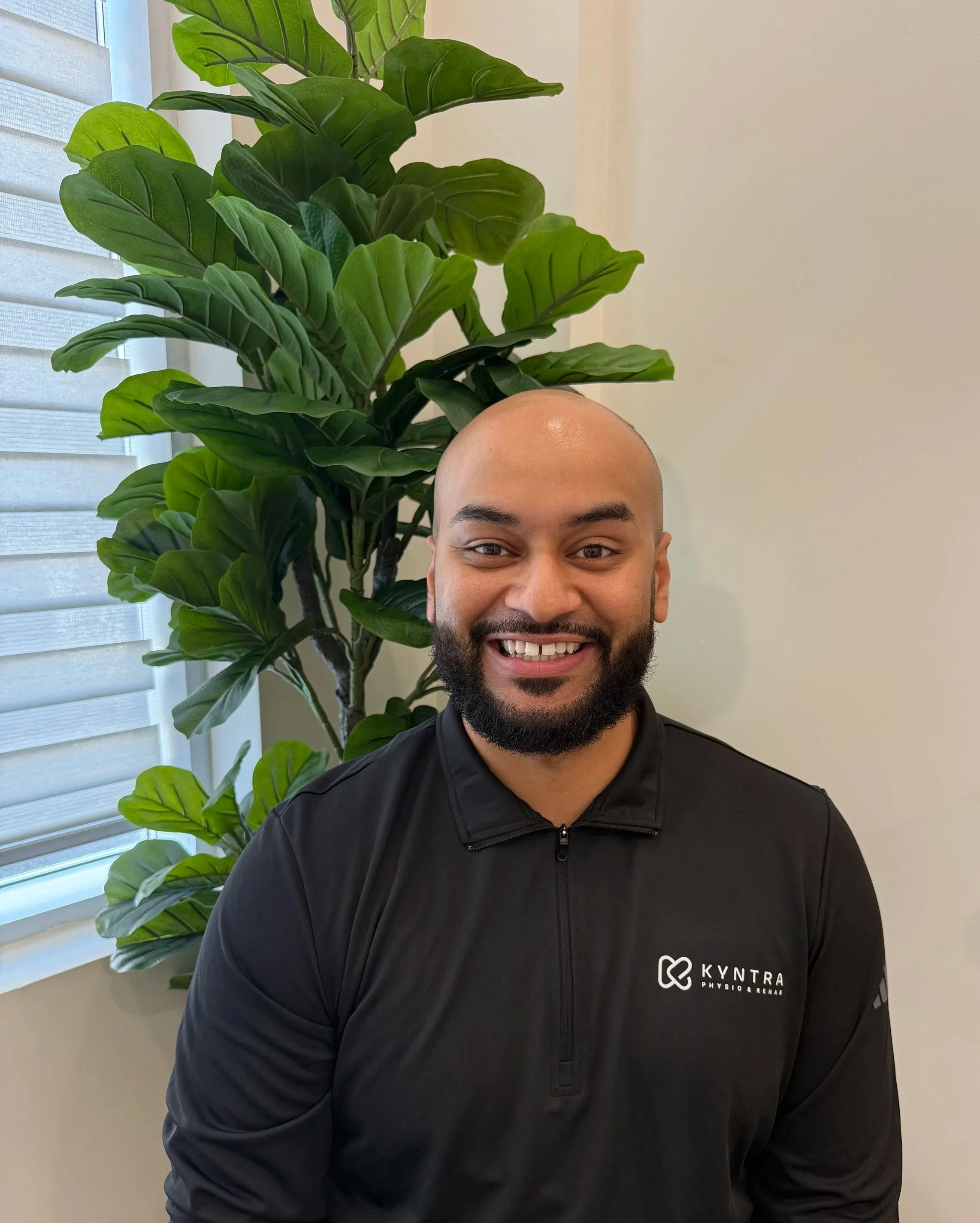 A smiling man with a beard and a shaved head wearing a black jacket with the logo of KYTERRA Physio & Rehab. Behind him is a large leafy green plant and window blinds.