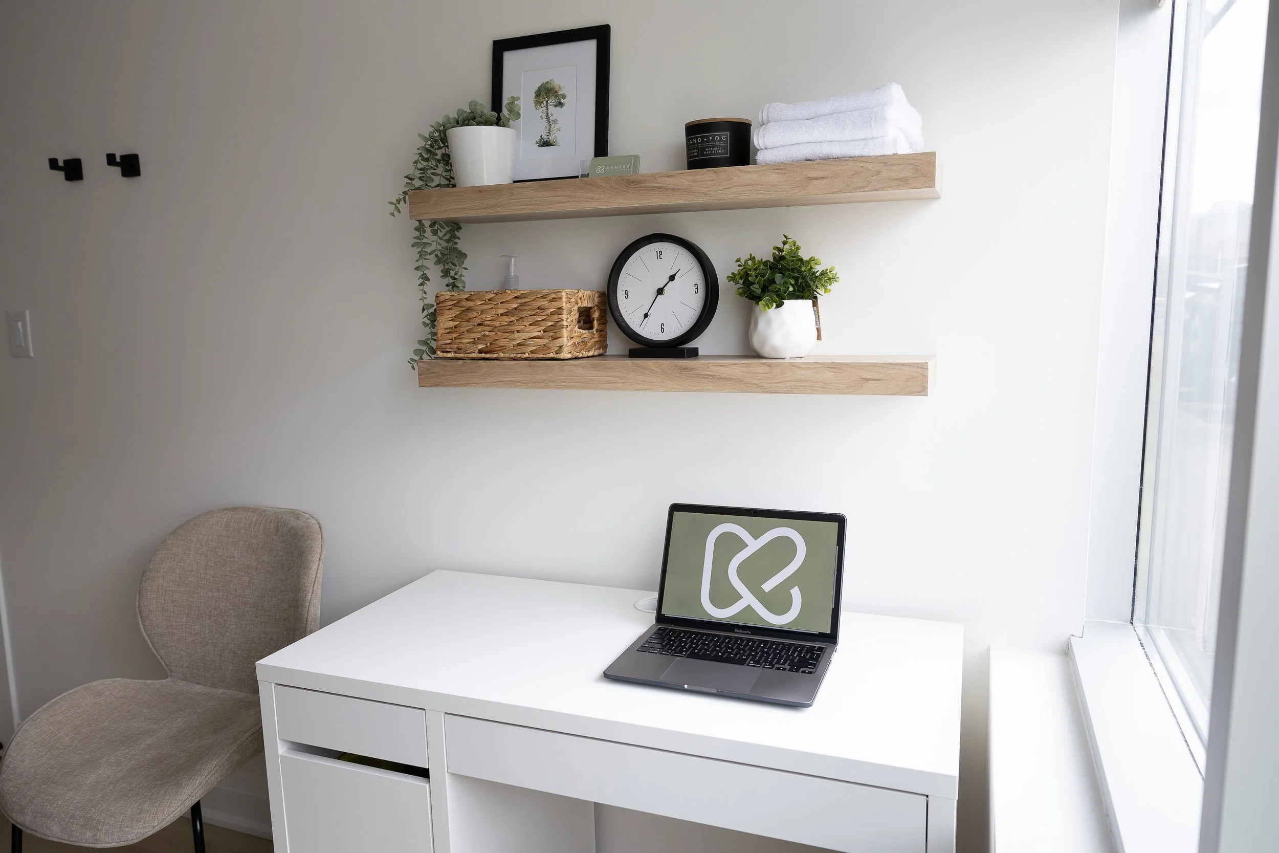 A modern workspace with a white desk, beige chair, and a MacBook displaying an abstract logo. Two wooden floating shelves on a white wall hold plants, a clock, neatly folded towels, and decorative items. A window illuminates the room with natural light.