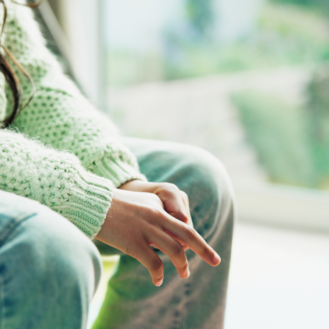 A person wearing a green knit sweater sitting near a window, with their hands resting on their lap.