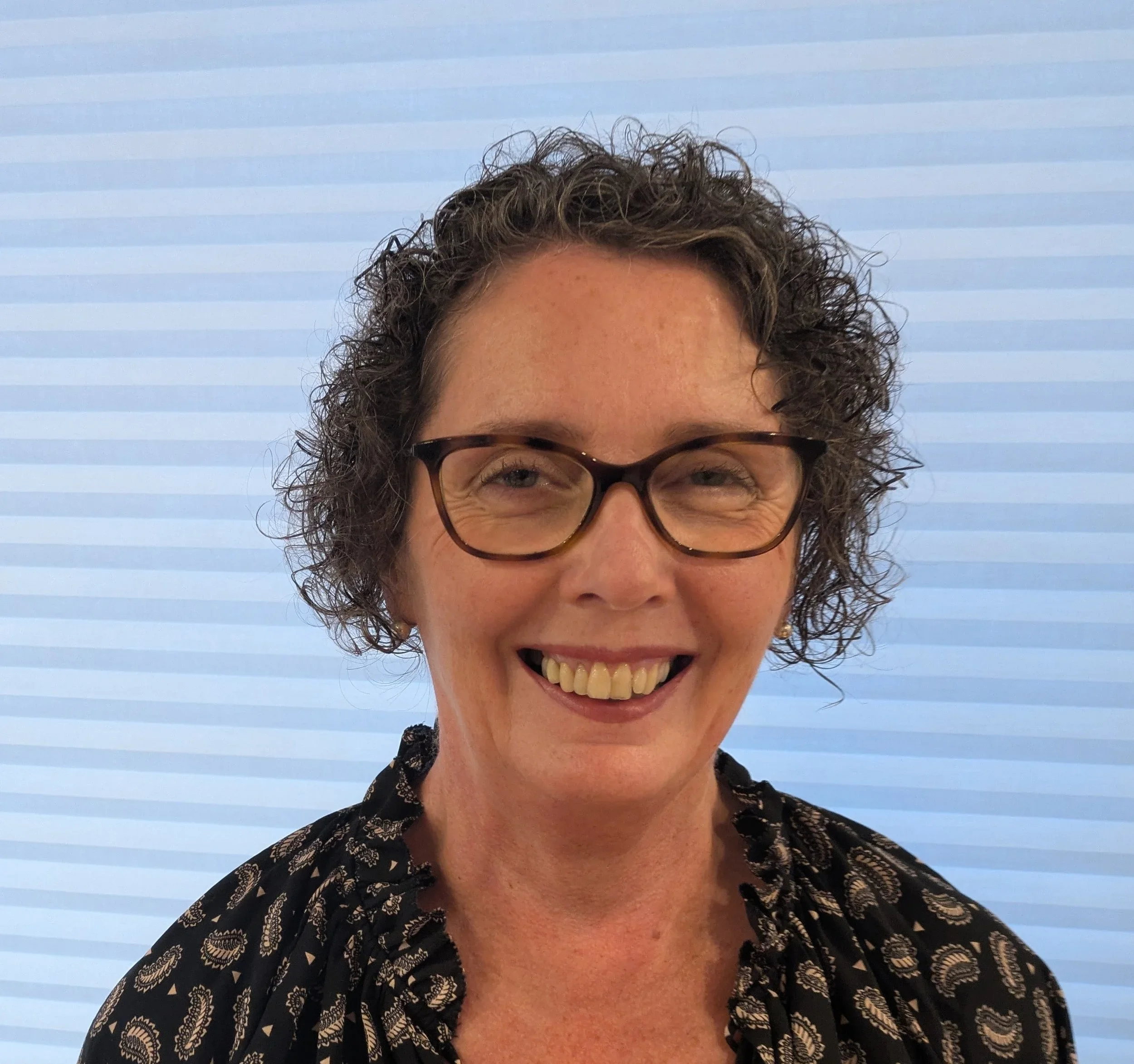 Close-up of a smiling woman with curly hair and glasses, wearing a dark patterned blouse, in front of a light blue and white striped background.