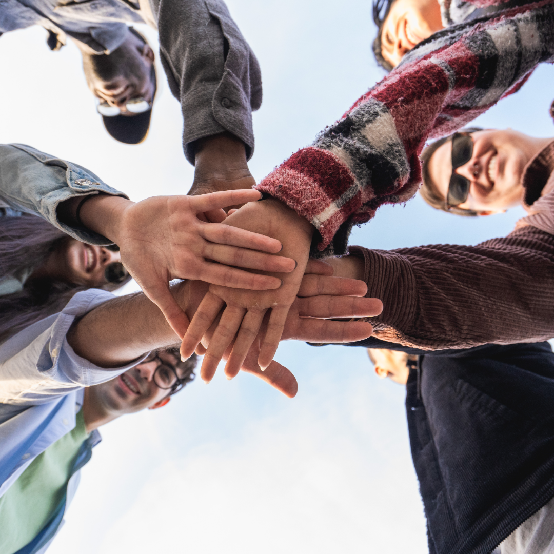 Group of young adults huddling together outdoors, stacking their hands in the center in a show of teamwork and friendship, with a clear sky in the background.