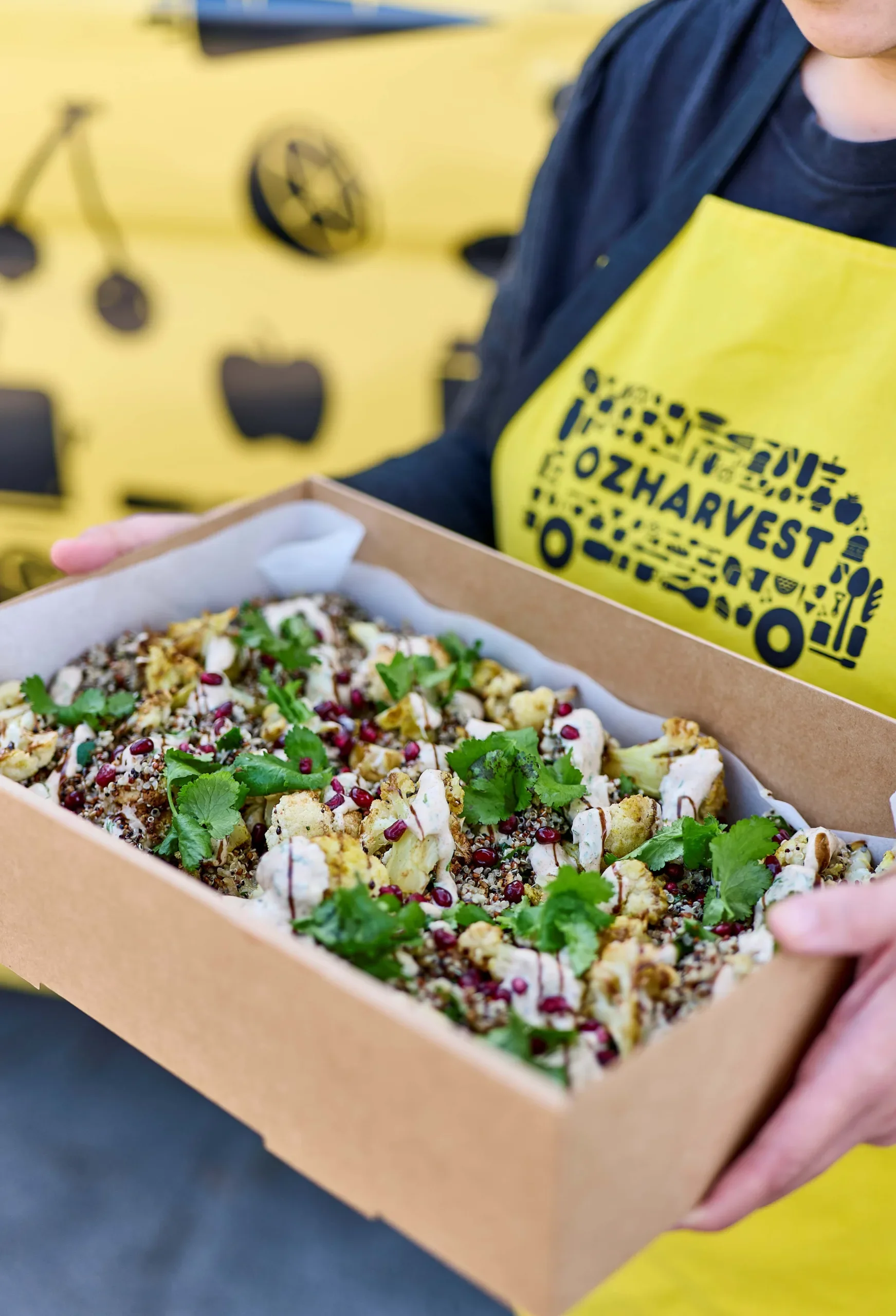 A person holding a cardboard box filled with a colorful salad topped with herbs and pomegranate seeds, with a person wearing a yellow apron that has a black graphic and text. In the background, there is a yellow wall with black illustrations of various objects, including glasses and a bicycle.