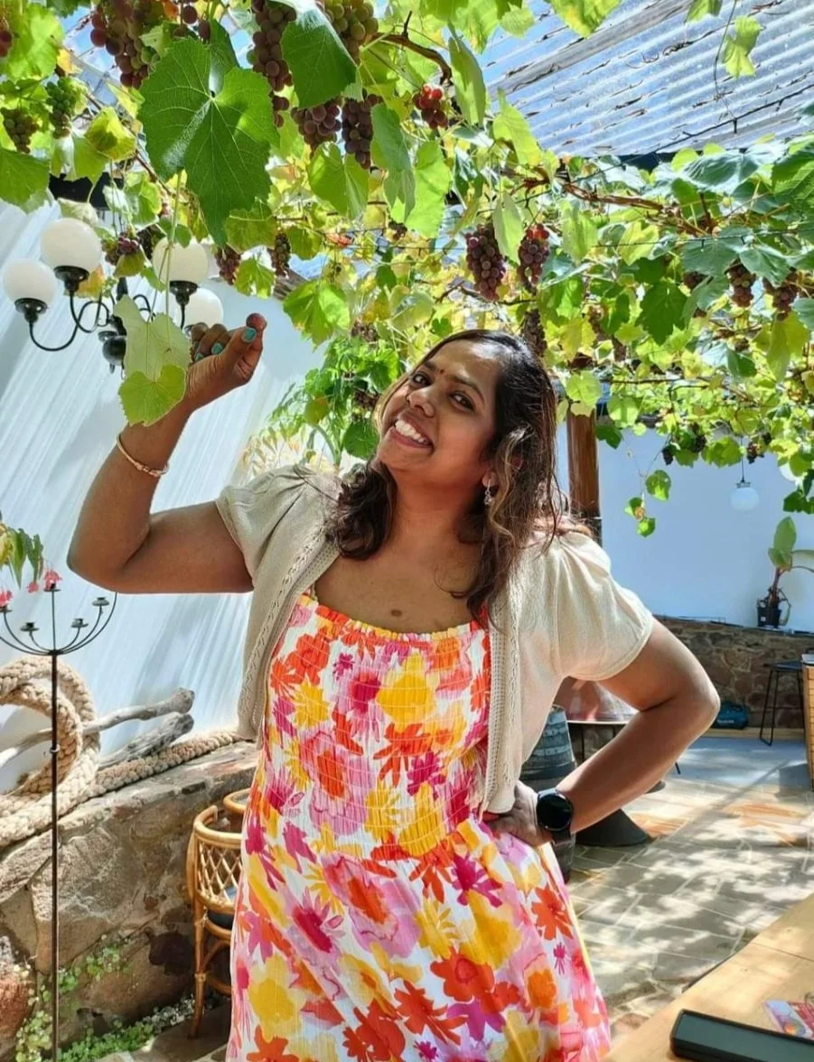 A woman smiling and holding a grape in a vineyard or patio with grapevines overhead, a stone wall, a lamp, and outdoor furniture.