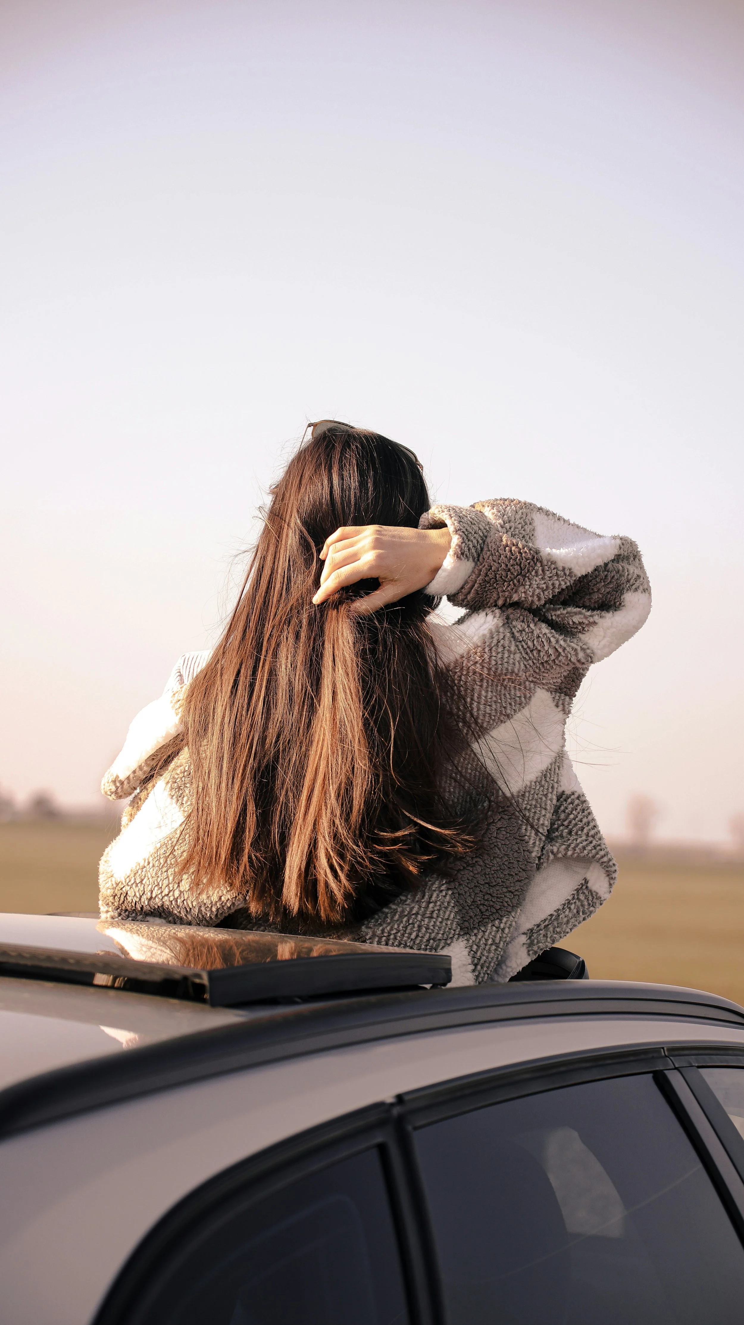 A woman with long brown hair wearing a beige and gray checkered coat is sitting on the roof of a car, holding her hair with one hand, against a clear sky background.
