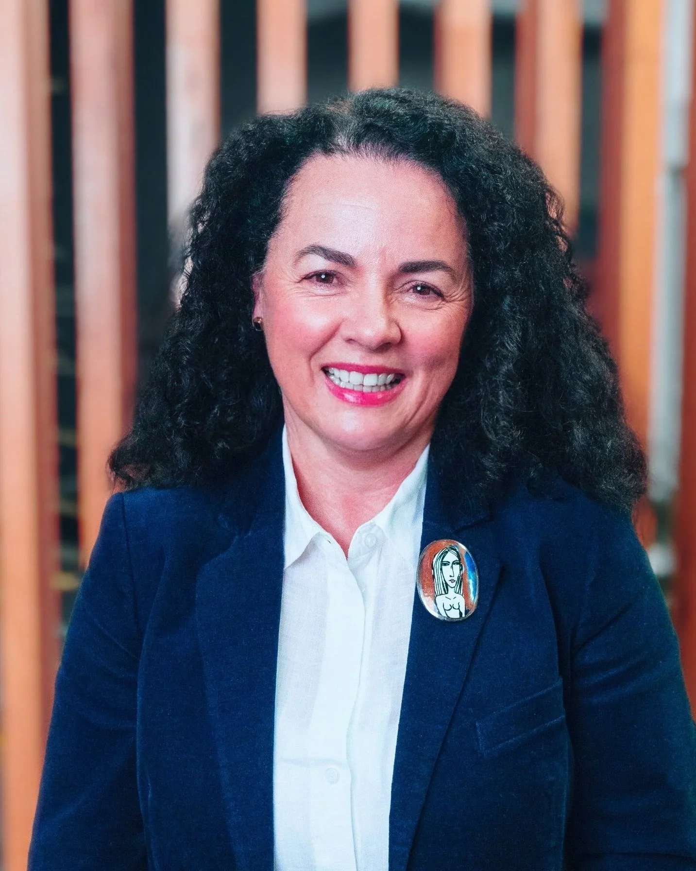 A woman with dark, curly hair smiling at the camera, dressed in a white shirt and dark blue blazer, with a colorful badge pin on her blazer, standing in front of a wooden slatted background.