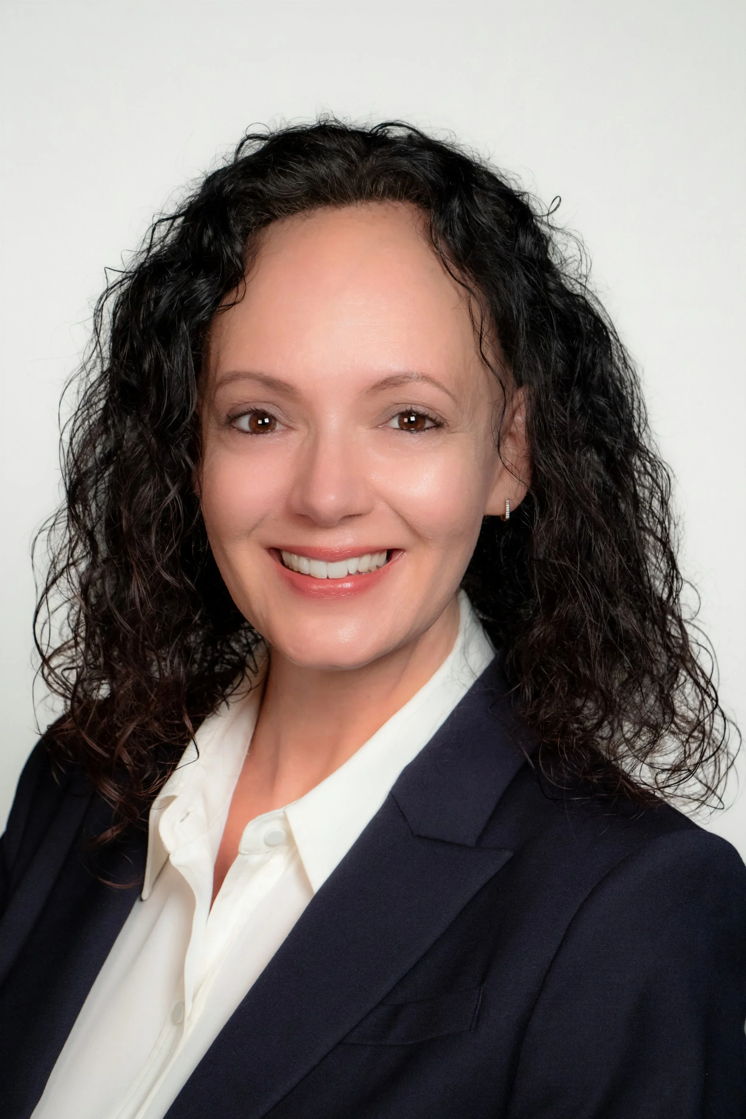 Woman with curly dark hair, wearing a dark blazer and white blouse, smiling against a plain white background.