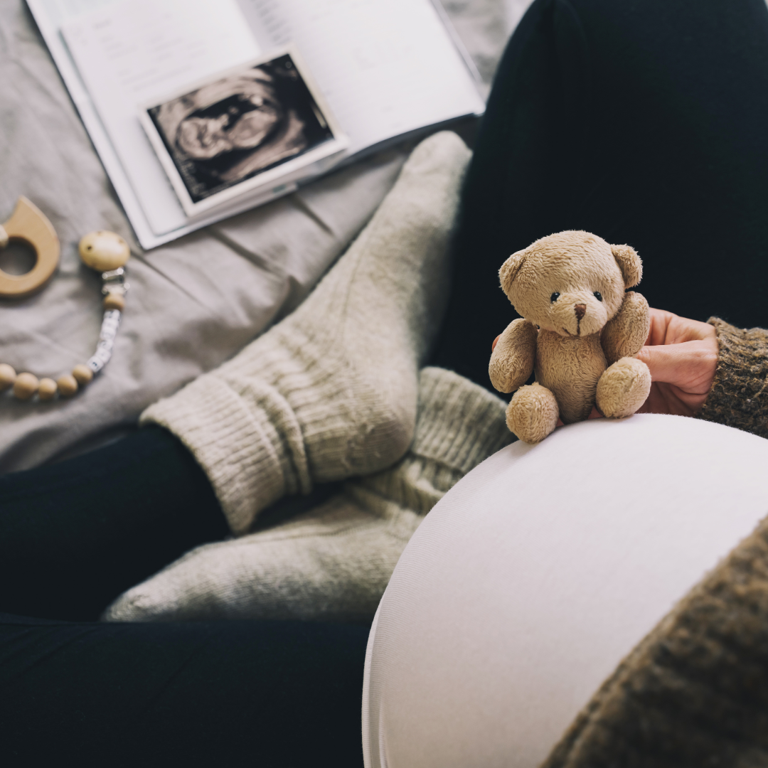 Pregnant woman holding a small teddy bear while sitting on a couch with a pregnancy book and a pacifier nearby.