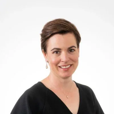 A woman with short brown hair smiling, wearing a black top and pearl earrings, against a plain white background.