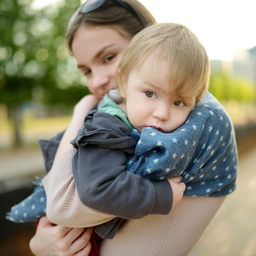 A woman holding a young boy with blond hair and blue eyes outdoors. The boy is resting his head on the woman's shoulder, and they are surrounded by greenery.