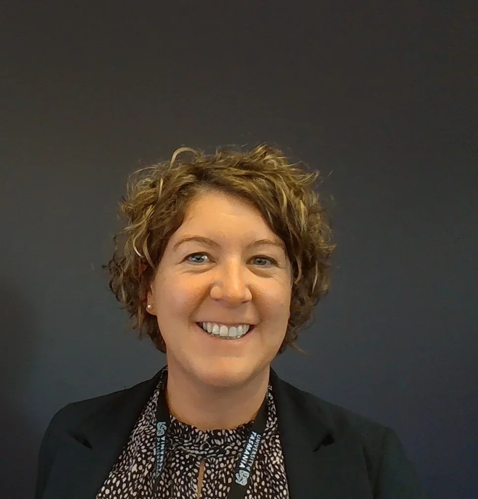 A woman with short, curly brown hair smiling at the camera against a dark gray background.
