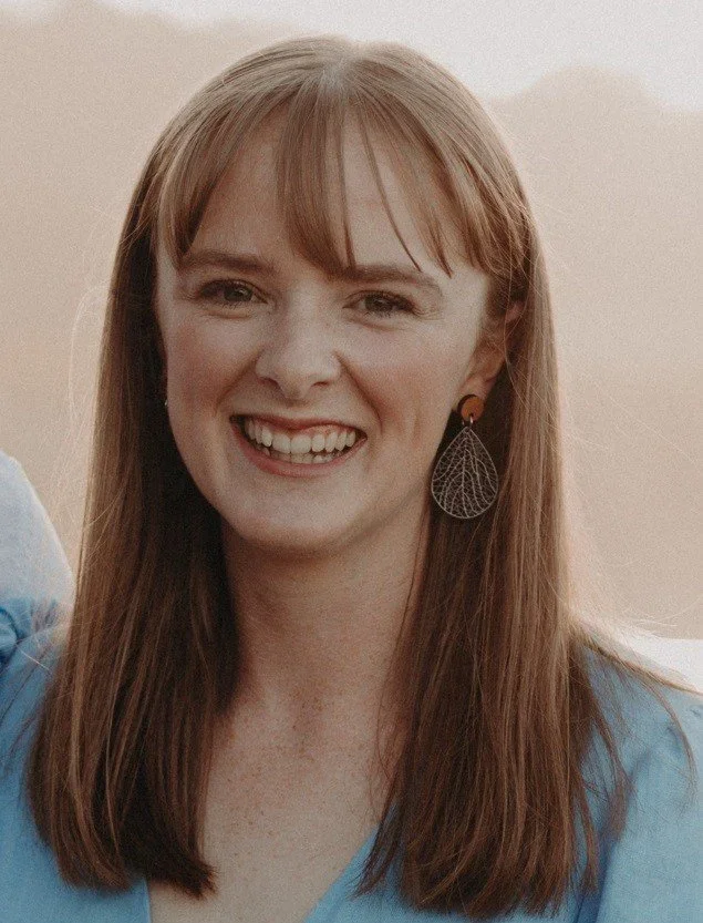 A young woman with long red hair smiling, wearing a blue top and leaf-shaped earrings.