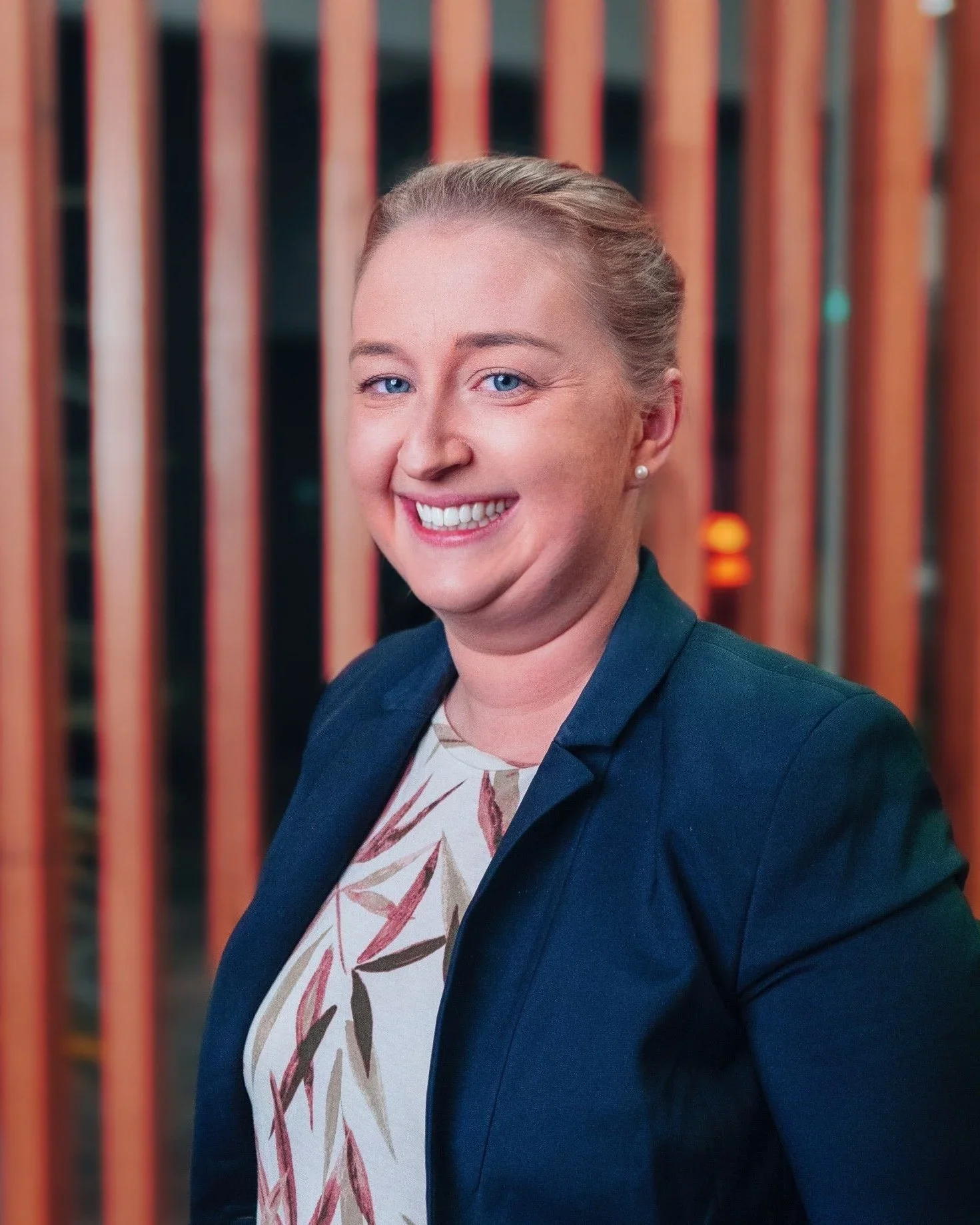 Headshot of a woman with blonde hair and blue eyes smiling, wearing a navy blazer and patterned blouse, standing in front of vertical wooden slats.