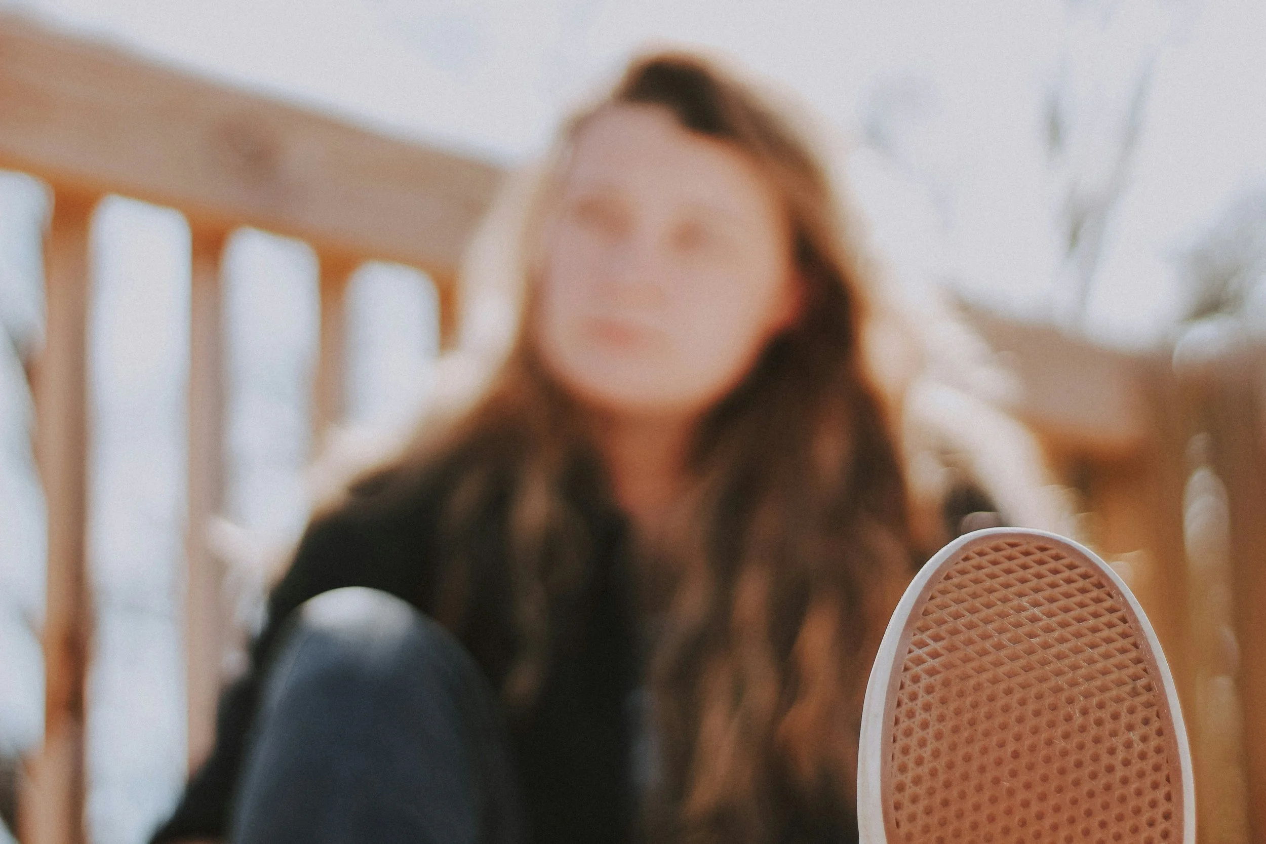 Blurred image of a person with long hair, sitting indoors near a white staircase, with a sneaker in the foreground.