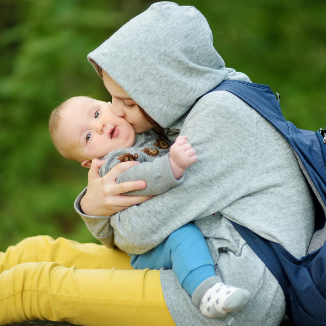 A person in a hoodie and yellow pants holding and kissing a baby outdoors.