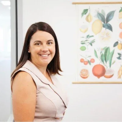 Smiling woman with dark hair standing in front of a botanical poster featuring various fruits and plants.