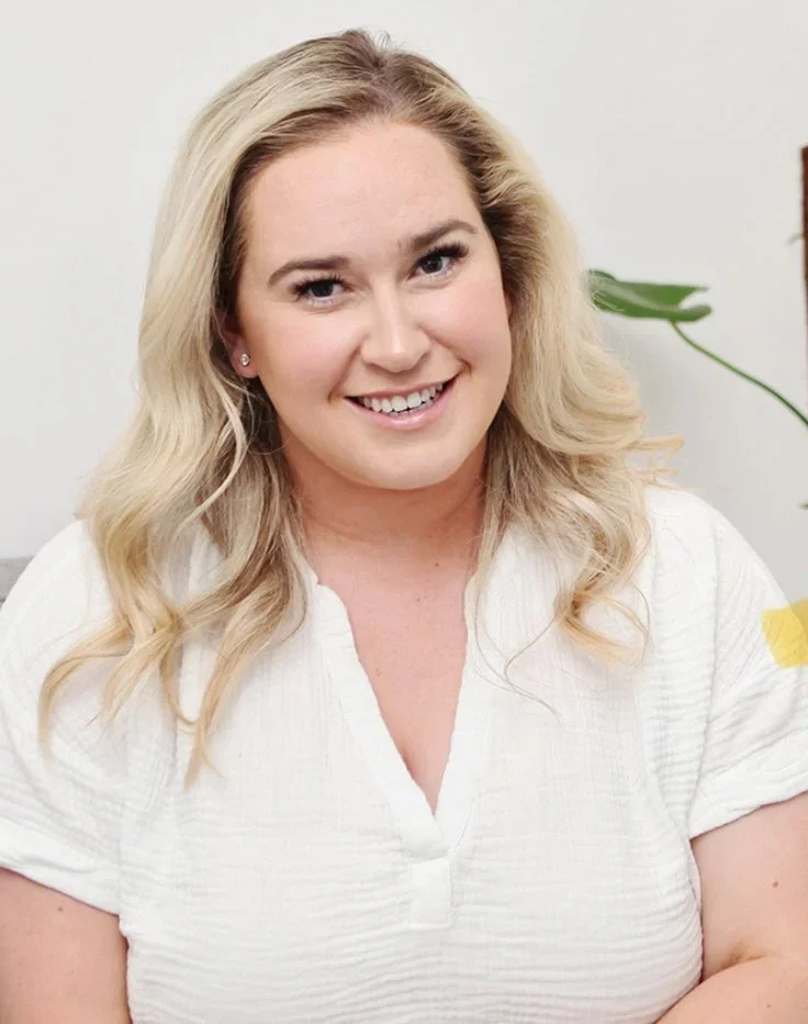 A smiling woman with blonde hair wearing a white top, sitting indoors near a plant.